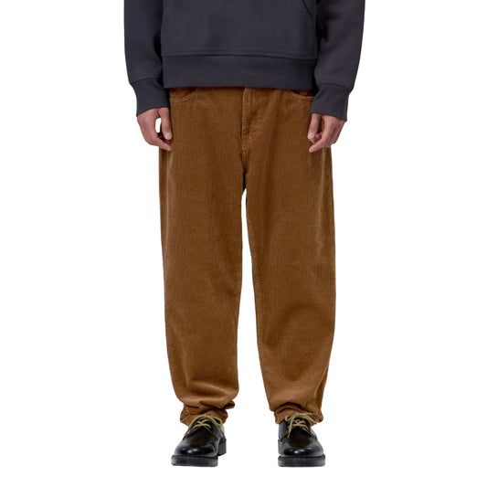 Calças Carhartt WIP Newel Pant em Hamilton Brown Rinsed — modelo masculino de frente em close ao corpo, detalhe do corte relaxado afunilado em bombazina Coventry de oito nervuras castanha Hamilton Brown rinsed, cintura regular, bolsos frontais, sapatos pretos de atacadores bege, fundo branco.