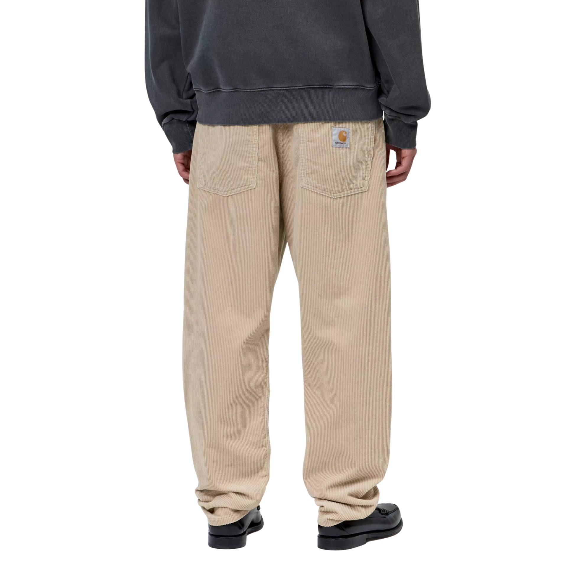 Calças Carhartt WIP Newel Pant em Wall Rinsed — modelo masculino de costas, meio corpo inferior, sweatshirt oversized cinzenta escura Carhartt WIP, calças em bombazina Coventry de oito nervuras bege Wall rinsed com corte relaxado afunilado, Square Label Carhartt WIP com "C" laranja no bolso traseiro direito, loafers pretos de bico redondo, fundo branco.