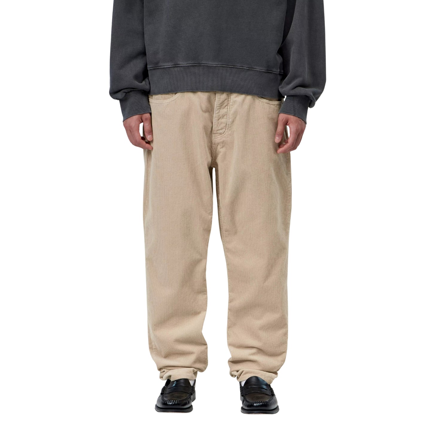 Calças Carhartt WIP Newel Pant em Wall Rinsed — modelo masculino de frente, meio corpo inferior, sweatshirt oversized cinzenta escura Carhartt WIP, calças em bombazina Coventry bege Wall rinsed com corte relaxado afunilado e cintura regular, bolsos frontais com rebites prateados visíveis, loafers pretos, fundo branco.