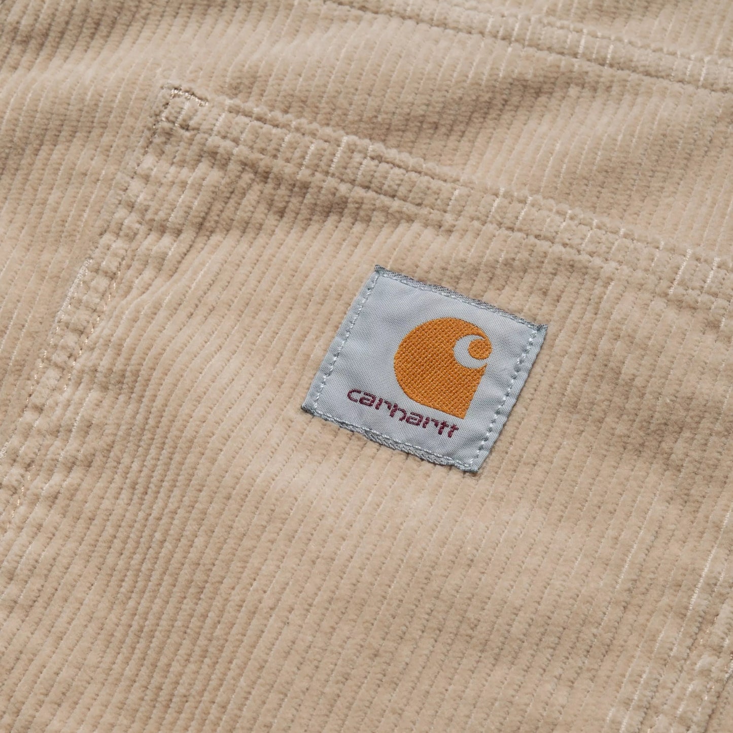 Calças Carhartt WIP Newel Pant em Wall Rinsed — detalhe do bolso traseiro esquerdo em close, bombazina Coventry de oito nervuras bege Wall rinsed, dois rebites prateados na abertura do bolso, textura canelada larga em destaque, fundo branco.