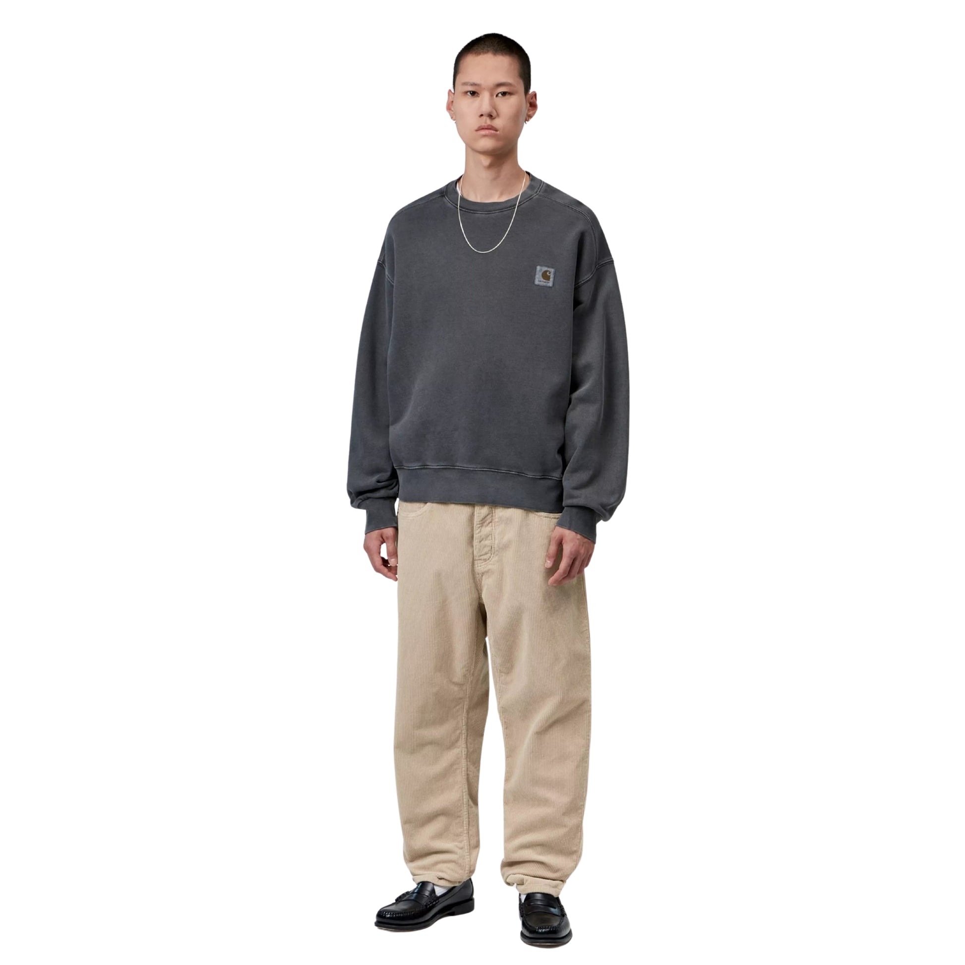 Calças Carhartt WIP Newel Pant em Wall Rinsed — modelo masculino de corpo inteiro, sweatshirt oversized cinzenta escura Carhartt WIP com Square Label no peito esquerdo, fio prateado ao pescoço, calças em bombazina bege Wall rinsed com corte relaxado afunilado a empilhar nos tornozelos, loafers pretos com meias brancas visíveis, fundo branco.