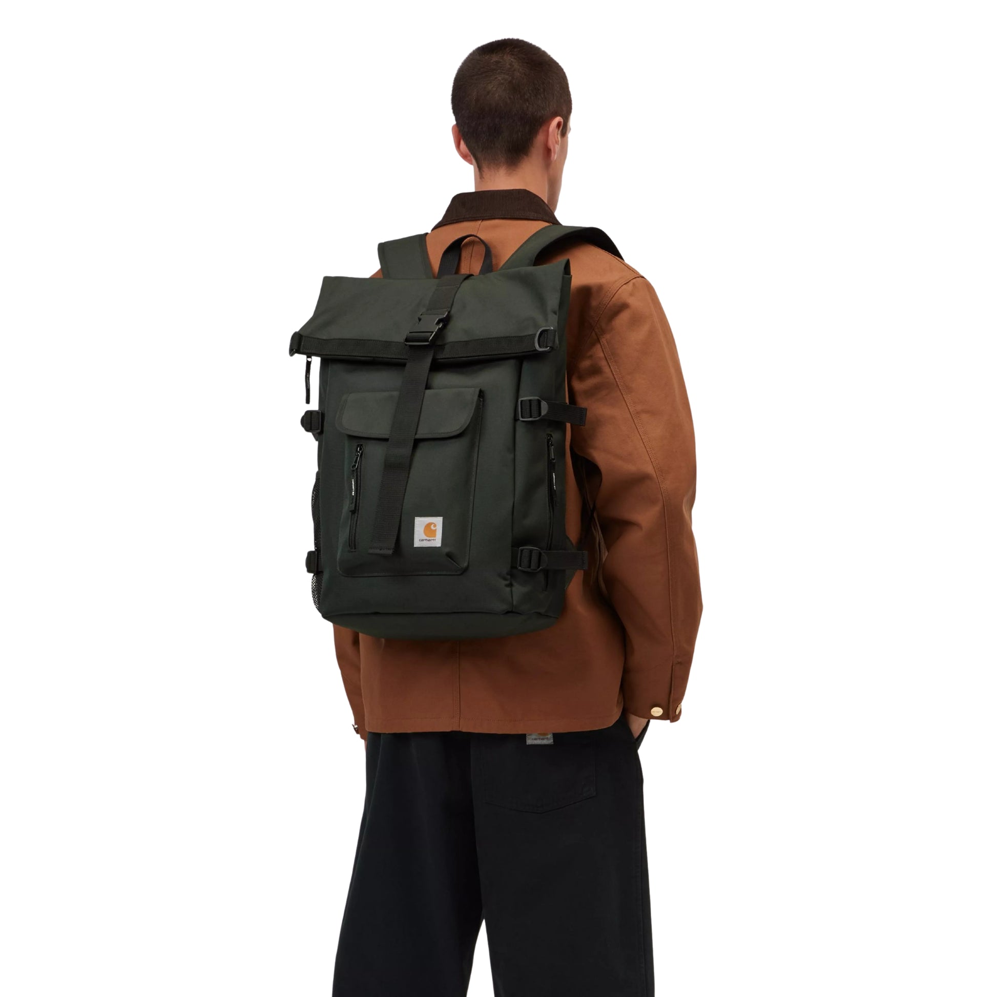 Mochila Carhartt WIP Philis Backpack em Raven — modelo de costas com mochila às costas, lapela com fecho, bolso frontal com fecho de correr, patch "C" Carhartt dourado e fivelas laterais visíveis, combinado com casaco Carhartt WIP castanho e calças largas pretas.