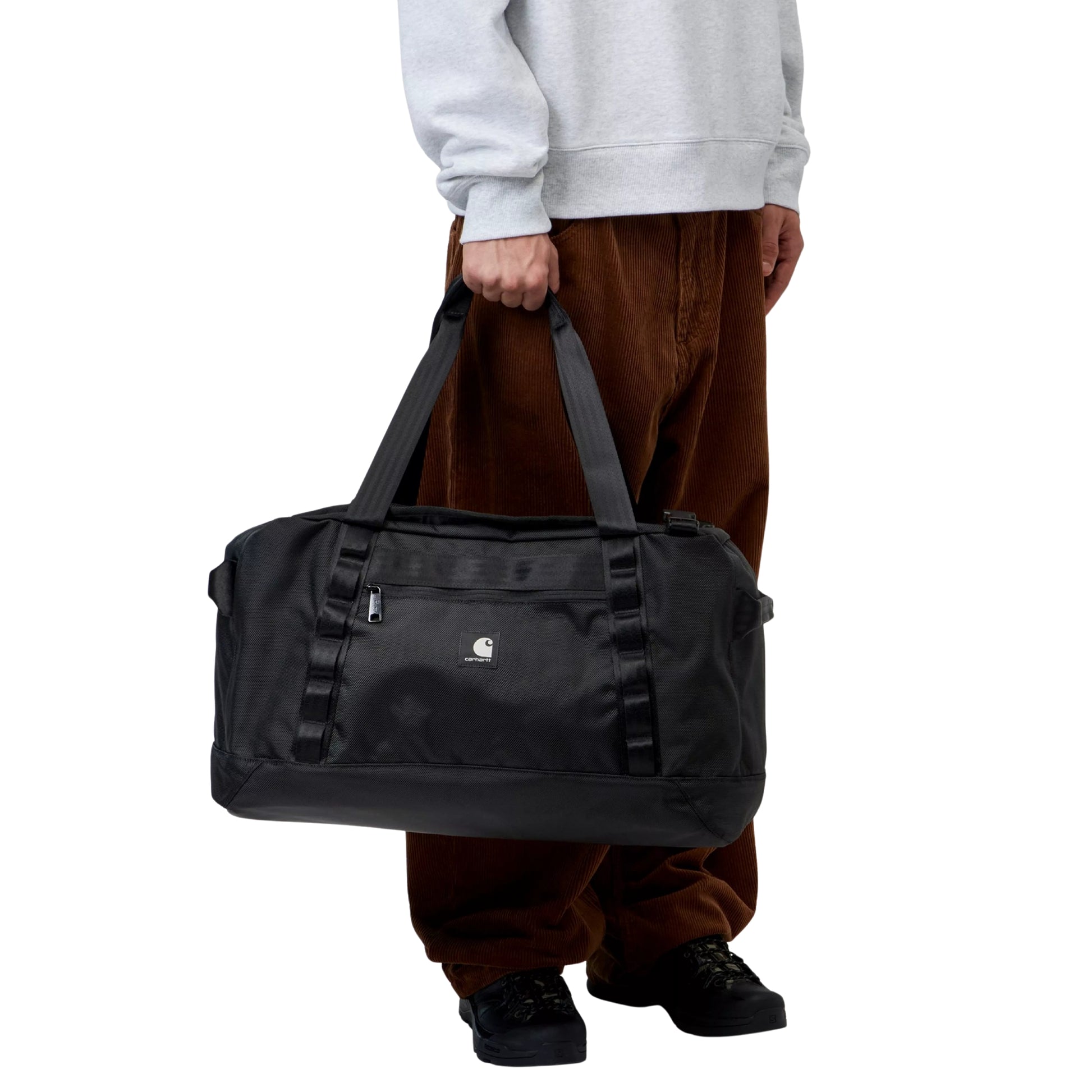 Mala de viagem Carhartt WIP Richmond Small Duffle Bag em Black — modelo masculino de meio corpo segurando a mala pelas duas pegas superiores em nylon preto, compartimento principal com fecho de correr e puxador prateado visível, bolso frontal com fecho de correr e etiqueta quadrada em tecido com logótipo "C" Carhartt branco, fivelas de compressão laterais pretas, calças wide-leg de bombazine castanha e sapatilhas pretas, nylon de textura pesada preto, fundo branco.