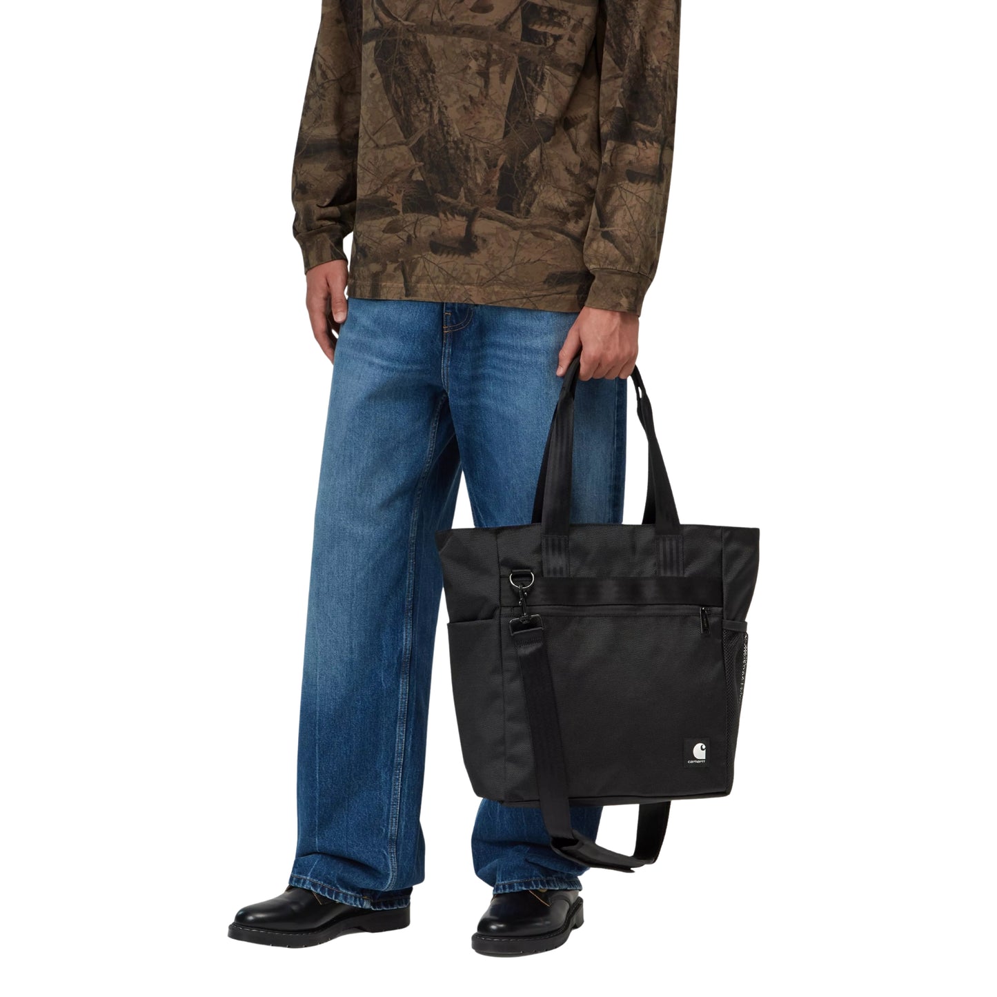 Tote bag Carhartt WIP Richmond em Black — modelo masculino de frente, meio corpo, camisa de manga comprida verde-militar com padrão camuflado, calças de ganga azul largas, sapatilhas pretas, a segurar a bolsa pela pega superior direita ao lado do corpo, logótipo Carhartt WIP branco visível na frente inferior, nylon texturado preto impermeável, fundo branco.