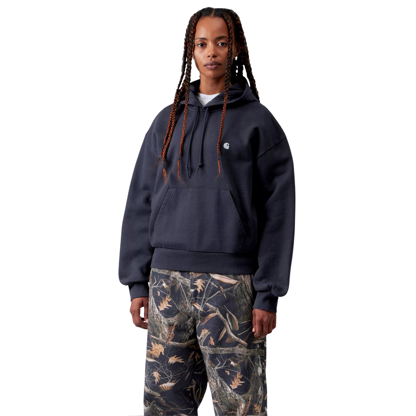 Camisola com capuz Carhartt WIP W' Hooded Casey Sweatshirt em Dark Navy — modelo feminino de frente, meio corpo superior, cabelo em tranças compridas soltas, camisola azul-marinho escura com corte balão e ombros caídos, capuz com cordão, bolso de canguru, logótipo "C" prateado no peito esquerdo, calças cargo wide-leg com print Realtree camo, fundo branco.