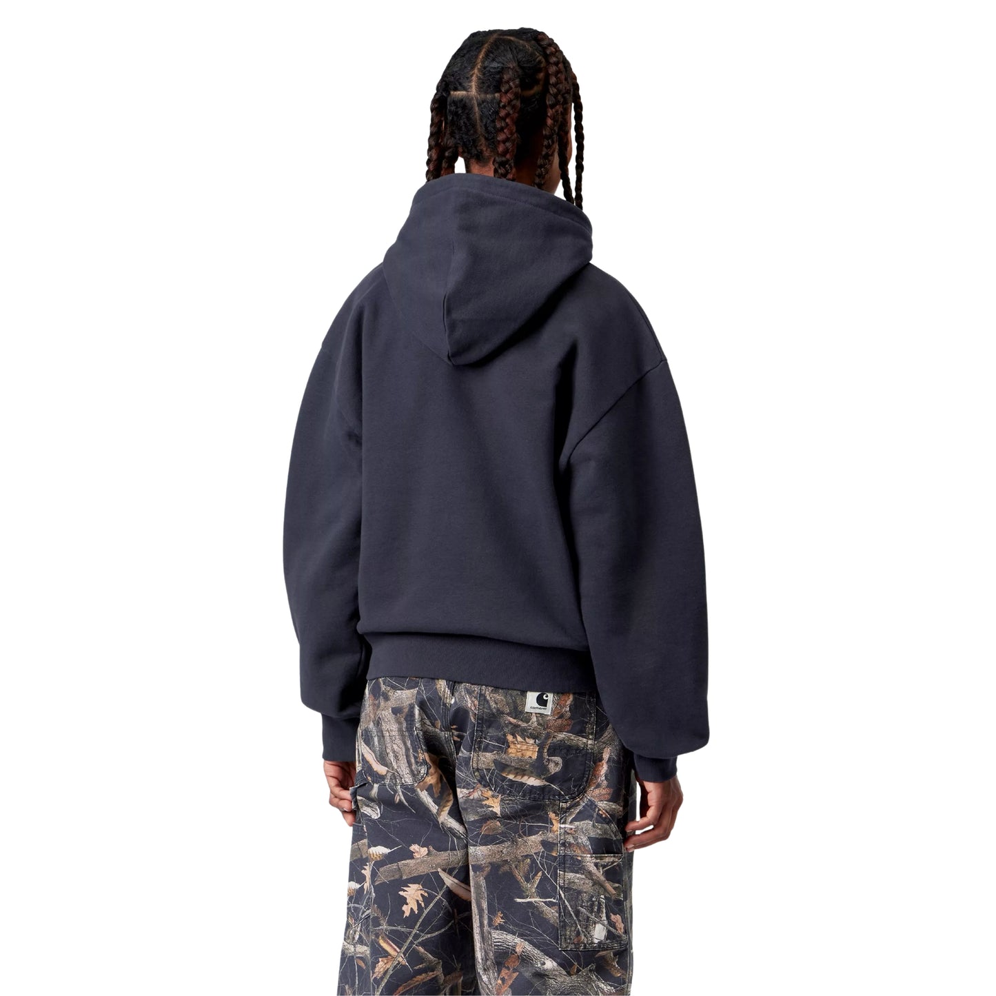 Camisola com capuz Carhartt WIP W' Hooded Casey Sweatshirt em Dark Navy — modelo feminino de costas, meio corpo superior, cabelo em tranças apanhadas, camisola azul-marinho escura com corte balão, ombros caídos bem pronunciados, capuz volumoso, barra canelada, calças cargo wide-leg com print Realtree camo e Square Label Carhartt WIP visível na cintura, fundo branco.