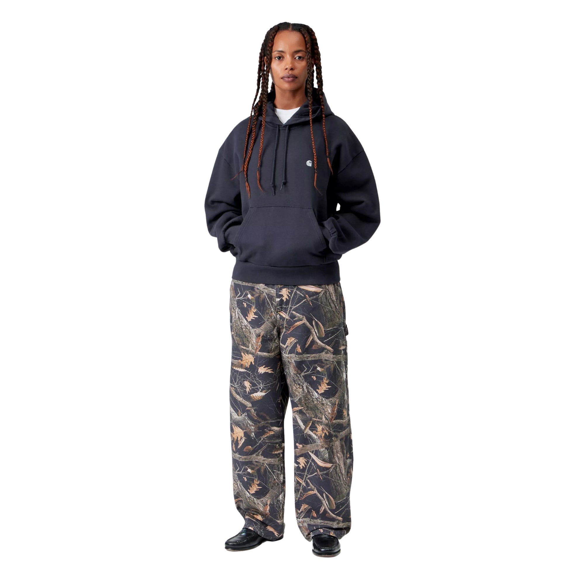 Camisola com capuz Carhartt WIP W' Hooded Casey Sweatshirt em Dark Navy — modelo feminino de frente, corpo inteiro, cabelo em tranças compridas soltas, camisola azul-marinho escura de corte balão com mãos no bolso de canguru, logótipo "C" prateado no peito esquerdo, calças cargo wide-leg com print Realtree camo, botas de bico fino pretas, fundo branco.