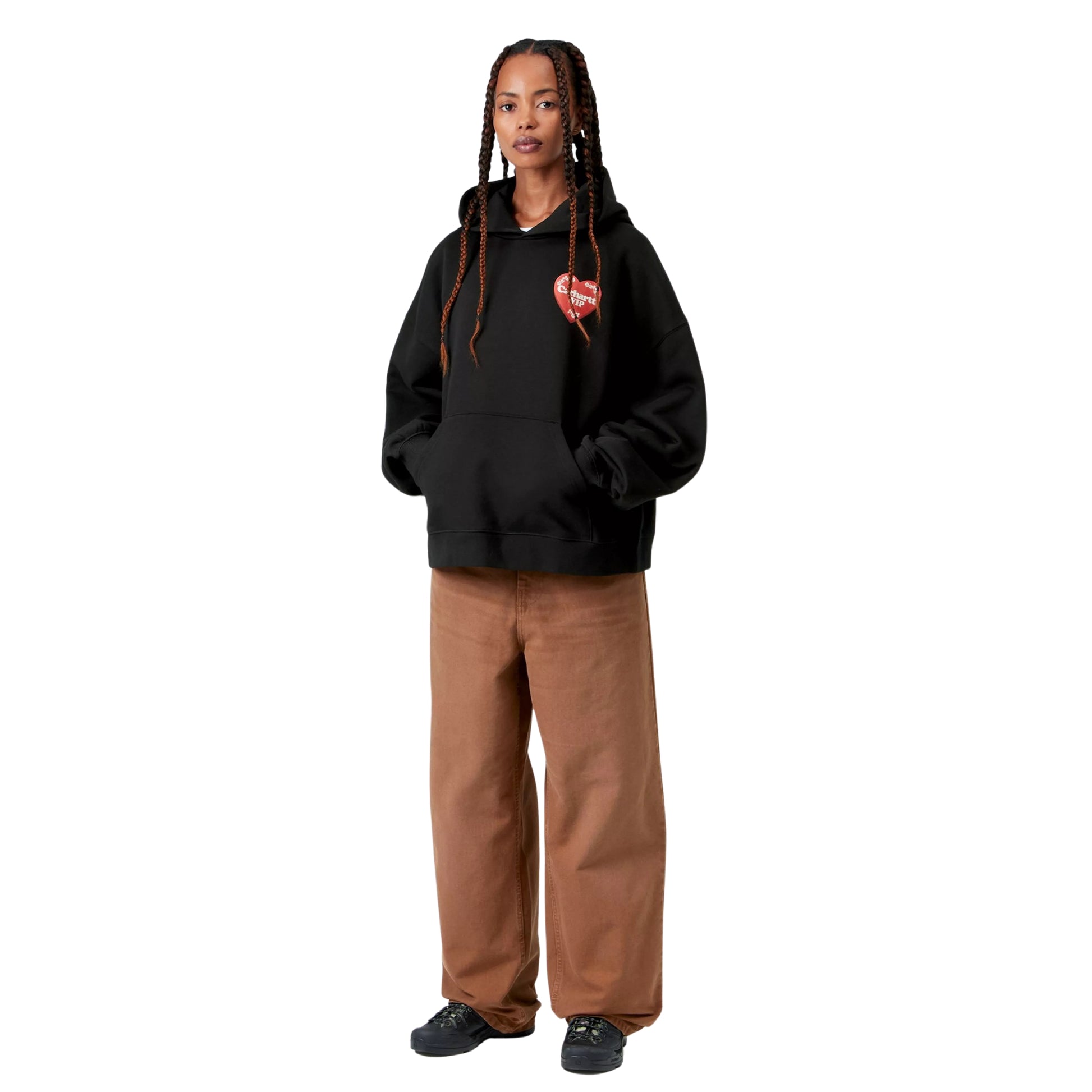 Camisola com capuz Carhartt WIP W' Hooded Hartt Case Sweat em Black — modelo feminino de frente, corpo inteiro, tranças longas, gráfico Hartt Case no peito esquerdo, bolso de canguru frontal, capuz volumoso, calças wide-leg cor de camel Carhartt WIP, sapatilhas pretas de sola grossa, malha de algodão escovada preta, fundo branco.