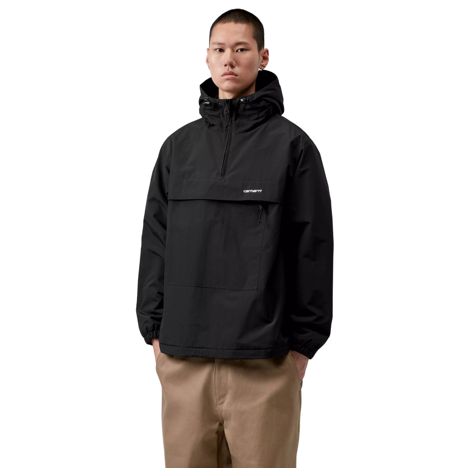Anorak Carhartt WIP Windbreaker Pullover em Black / White — modelo masculino de frente, meio corpo, cabelo curto escuro, brinco pequeno, capuz levantado com cordão e dois ajustadores pretos, fecho de correr a meio do peito, logótipo Script "carhartt." bordado à esquerda do peito em White, bolso frontal com fecho de correr e puxador preto visível, mãos nos bolsos, punhos elásticos, calças wide-leg bege Carhartt WIP, nylon Supplex® preto Black, fundo branco.
