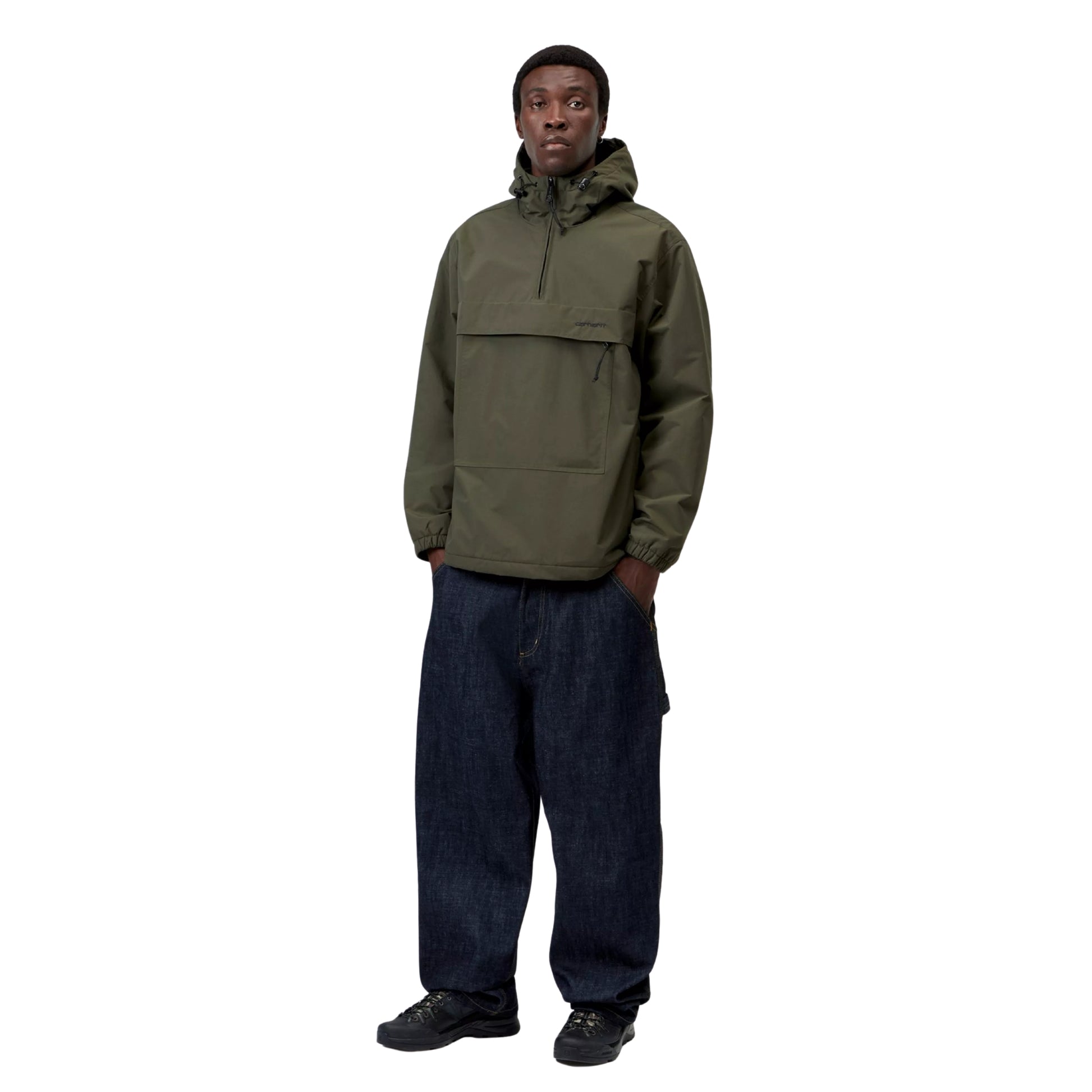 Anorak Carhartt WIP Windbreaker Pullover em Cypress / Black — modelo masculino de frente, corpo inteiro, gorro preto, capuz levantado, fecho de correr a meio do peito em Black, bolso frontal com fecho, mãos nos bolsos laterais, calças cargo denim azul-escuro largas, sapatilhas de trail pretas, nylon Supplex® verde militar Cypress, fundo branco.