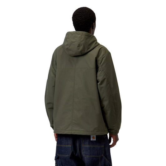 Anorak Carhartt WIP Windbreaker Pullover em Cypress / Black — modelo masculino de costas, gorro preto, capuz volumoso em Cypress, costas lisas sem gráfico com costuras estruturais visíveis, punhos elásticos, bainha com cordão de ajuste preto, etiqueta Carhartt WIP laranja na cintura das calças cargo denim azul-escuro largas, nylon Supplex® verde militar Cypress, fundo branco.