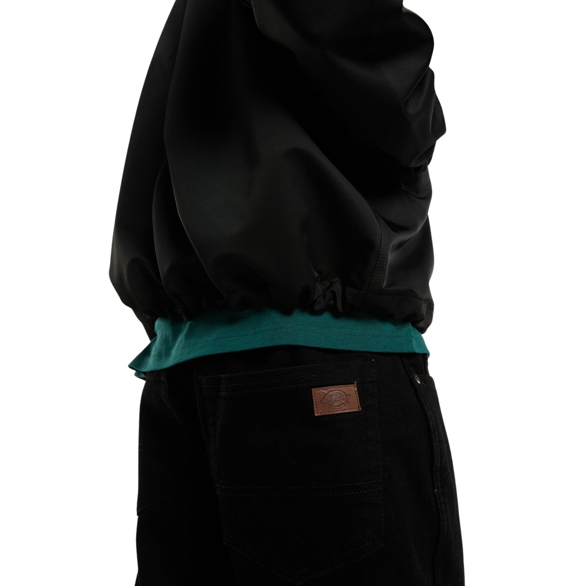 Casaco coach Dickies Parks em Preto, pormenor lateral da parte inferior, mostrando a manga e a bainha franzida sobre uma t-shirt verde.