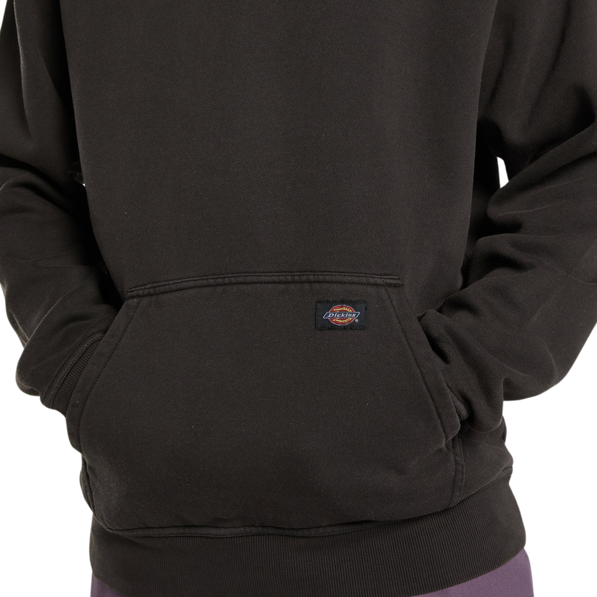 Sweat com carapuço Dickies Sutcliffe em Preto, close-up do bolso canguru com as mãos do modelo no interior, etiqueta tecida Dickies e bainha canelada.