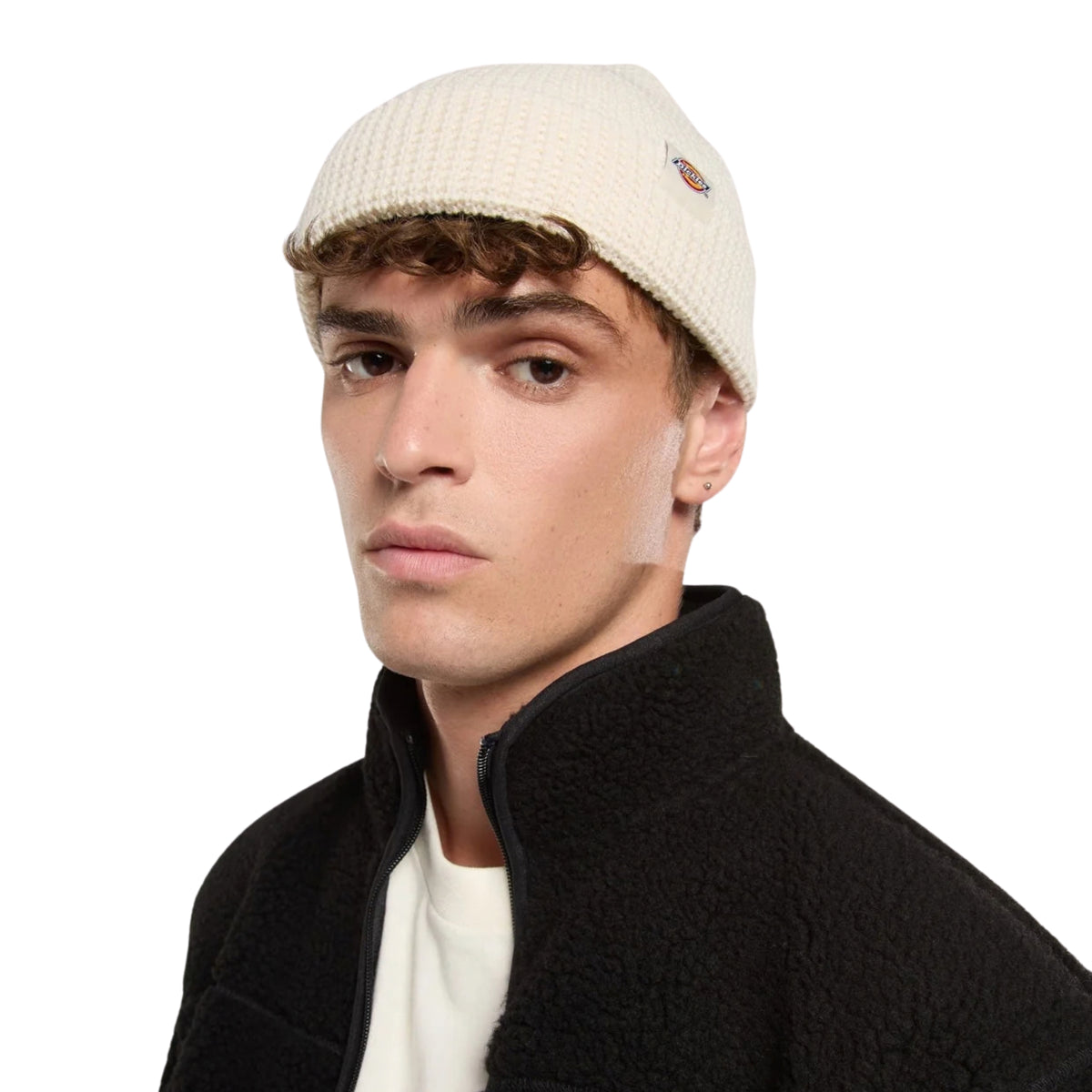 Gorro unissexo Dickies Woodworth Waffle Beanie em Ecru — modelo masculino em plano aproximado com patch bordada Dickies na dobra frontal, vestindo casaco polar preto.