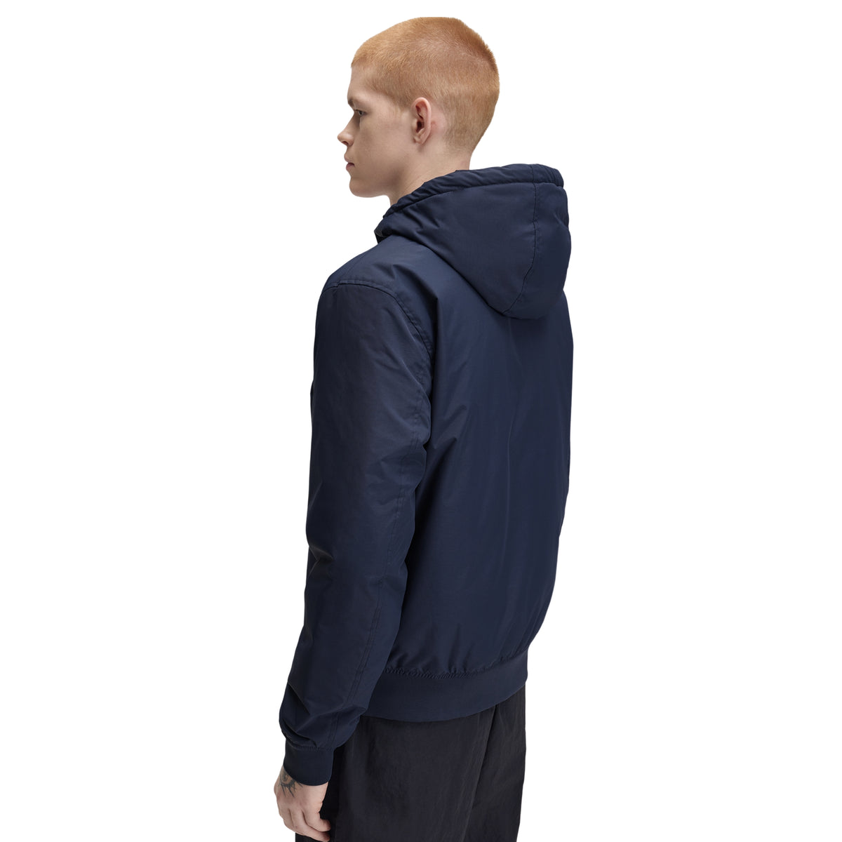 Casaco com carapuço para homem Fred Perry Hooded Brentham Jacket em Dark Airforce — modelo masculino de perfil direito visto por trás, corpo inteiro, casaco azul-marinha escuro com carapuço fechado, barra elástica canelada, regular fit, calças pretas, fundo branco.