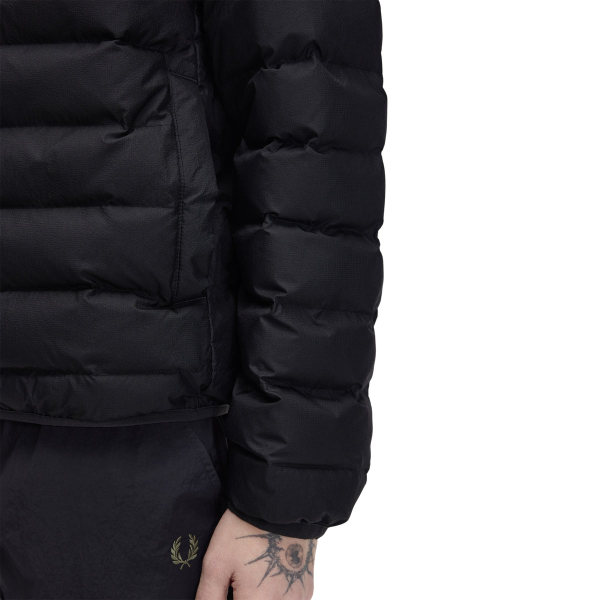 Casaco acolchoado para homem Fred Perry Insulated Jacket em Black — detalhe lateral direito, meio corpo, nylon reciclado preto, enchimento acolchoado horizontal em close-up, manga acolchoada com punho elástico, bolso lateral na costura visível, fundo branco.