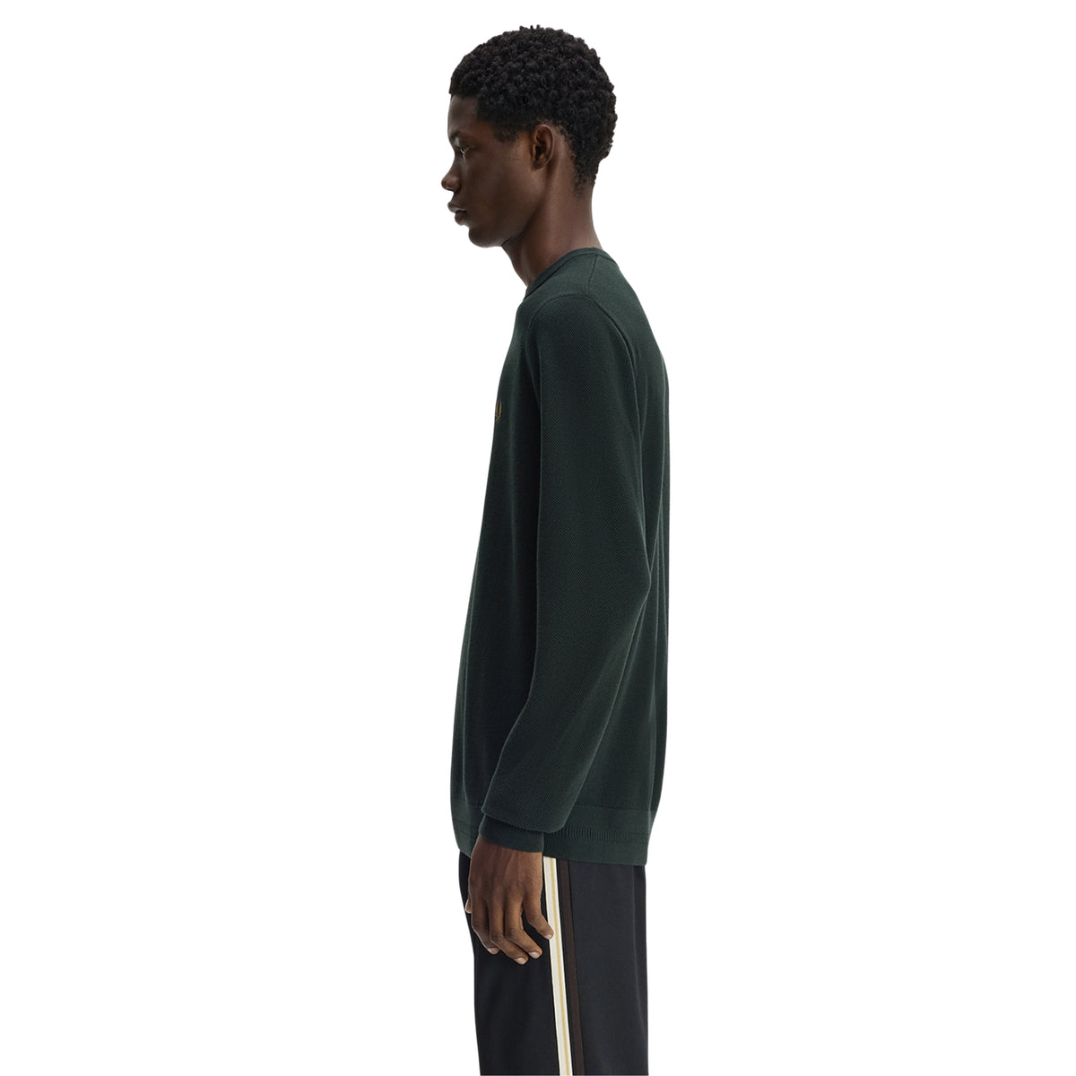 Camisola de malha para homem Fred Perry Pique Stitch Jumper em Night Green — modelo masculino de perfil direito, corpo inteiro, malha piqué verde-escuro Night Green, gola redonda canelada, punho canelado visível, regular fit, calças escuras com riscas laterais creme, fundo branco.