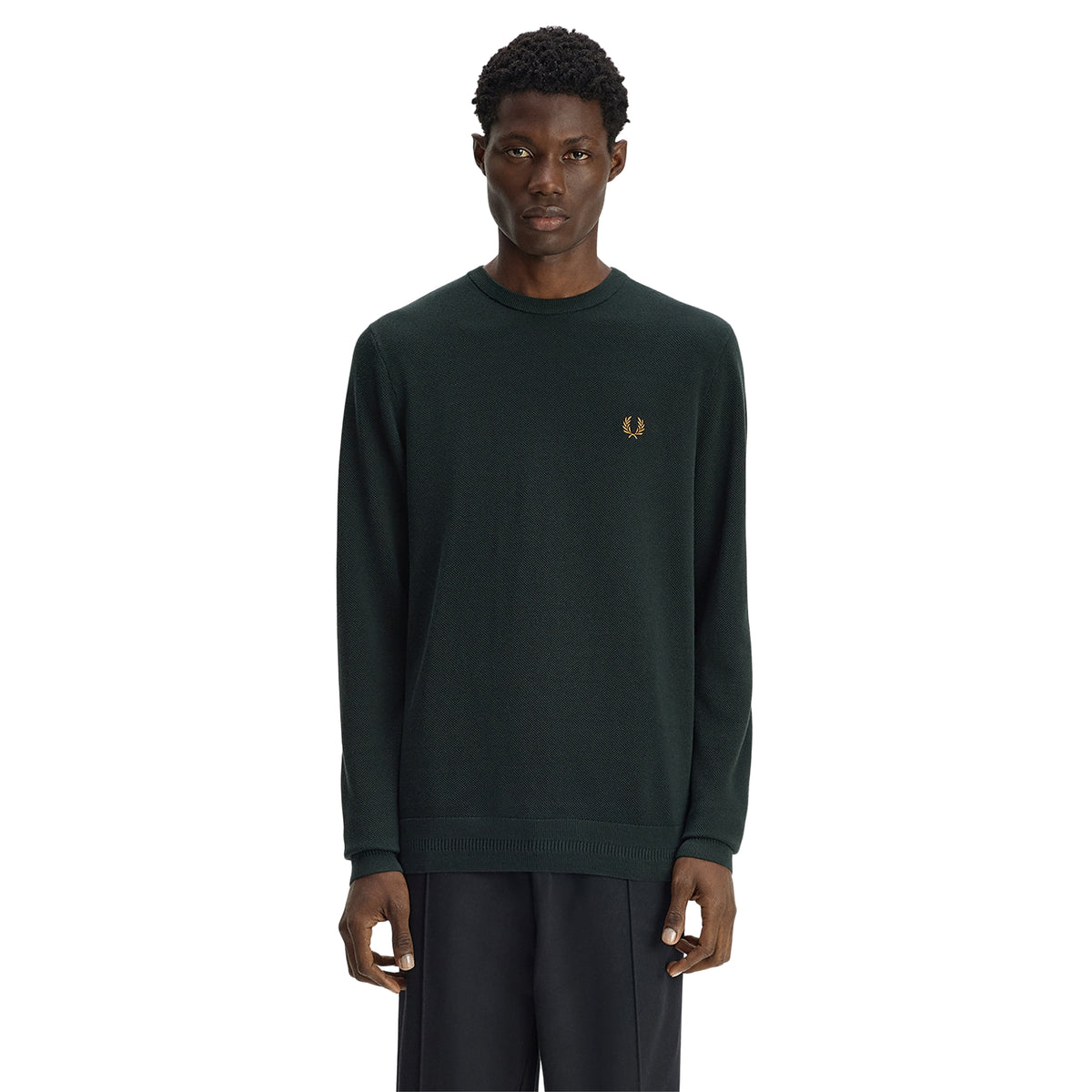 Camisola de malha para homem Fred Perry Pique Stitch Jumper em Night Green — modelo masculino de frente, corpo inteiro, malha piqué verde-escuro Night Green, gola redonda canelada, logo laurel wreath bordado a dourado no peito esquerdo, barra canelada, regular fit, calças pretas, fundo branco.