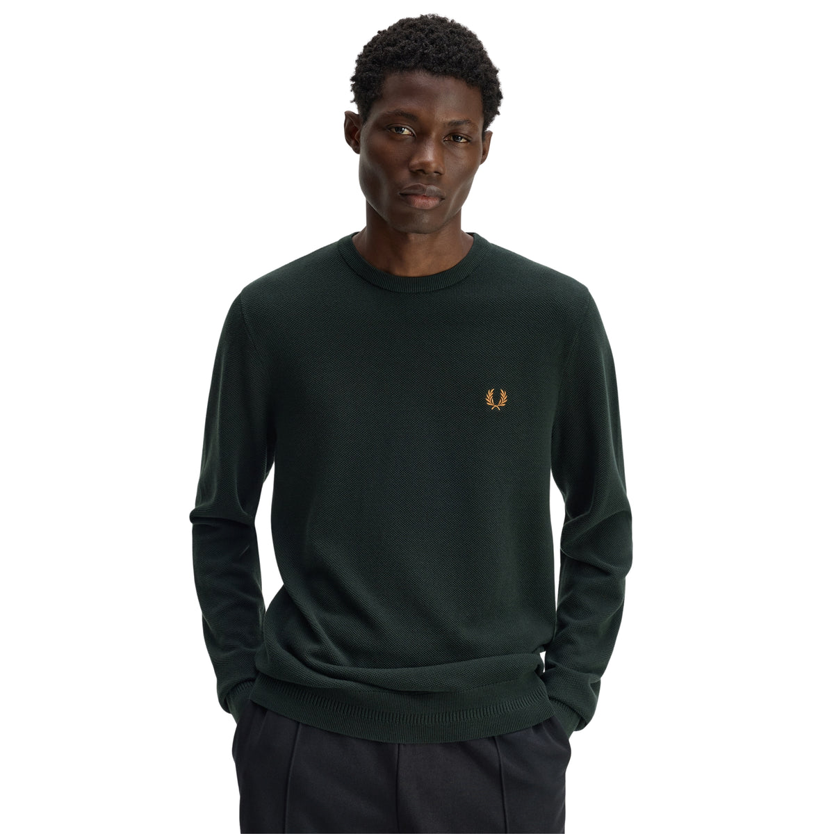 Camisola de malha para homem Fred Perry Pique Stitch Jumper em Night Green — modelo masculino de frente, meio corpo, malha piqué verde-escuro Night Green, gola redonda canelada, logo laurel wreath bordado a dourado no peito esquerdo, textura piqué visível, regular fit, mãos nas algibeiras das calças pretas, fundo branco.