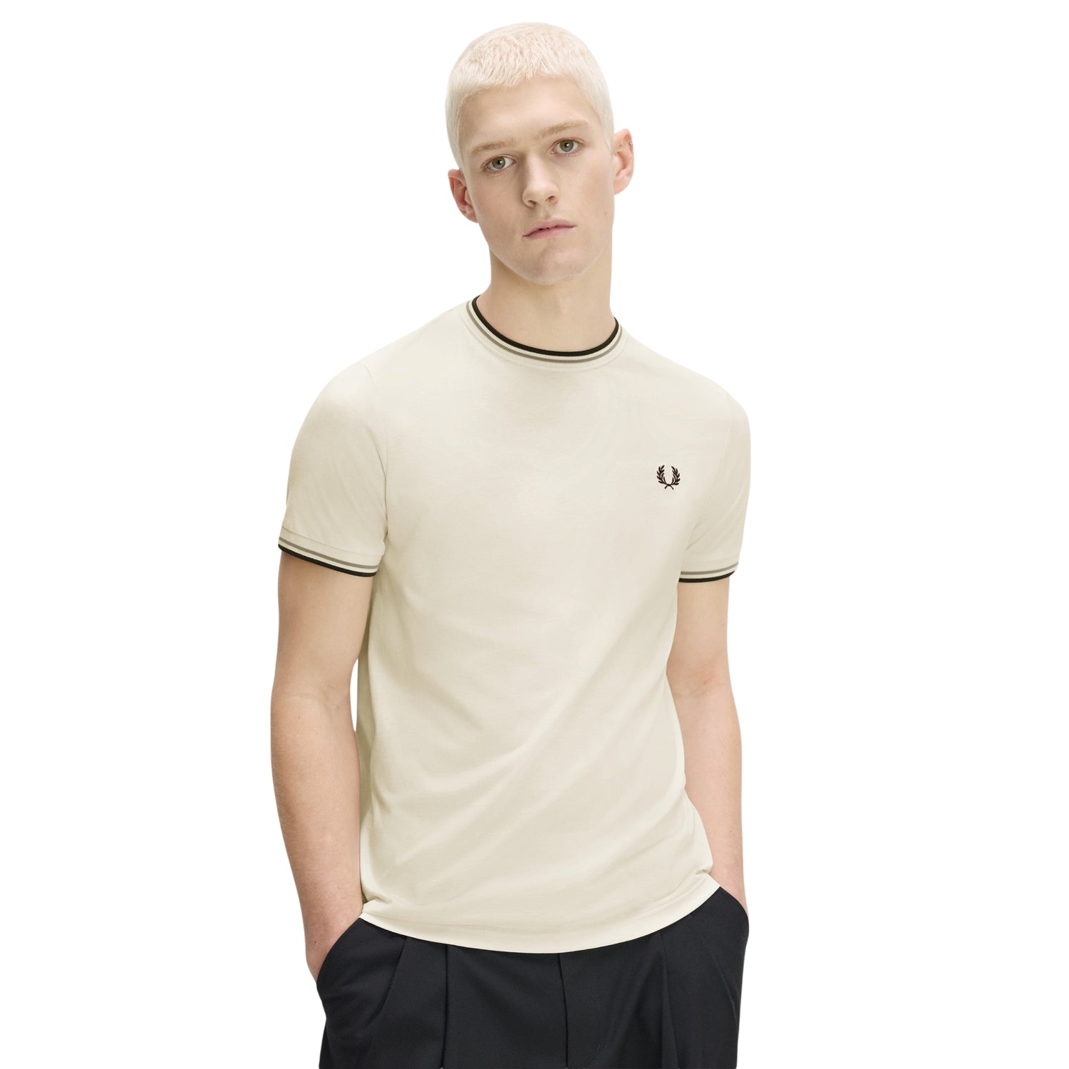 T-shirt Fred Perry Twin Tipped T-Shirt em Ecru — vista frontal com modelo, meio corpo, mãos nos bolsos, bordado Laurel Wreath em Dark Airforce no peito esquerdo, twin tipping Seagrass/Dark Airforce no colarinho e punhos, combinado com calças pretas.
