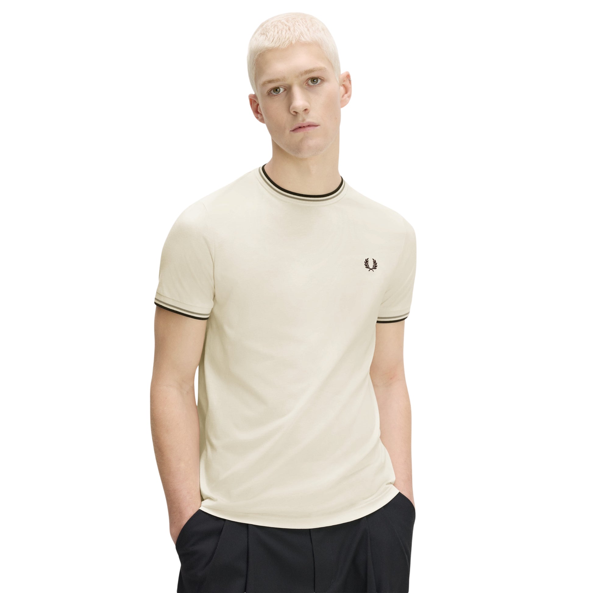 T-shirt Fred Perry Twin Tipped T-Shirt em Ecru — vista frontal com modelo, meio corpo, mãos nos bolsos, bordado Laurel Wreath em Dark Airforce no peito esquerdo, twin tipping Seagrass/Dark Airforce no colarinho e punhos, combinado com calças pretas.