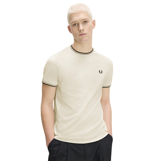 T-shirt Fred Perry Twin Tipped T-Shirt em Ecru — vista frontal com modelo, meio corpo, mãos nos bolsos, bordado Laurel Wreath em Dark Airforce no peito esquerdo, twin tipping Seagrass/Dark Airforce no colarinho e punhos, combinado com calças pretas.
