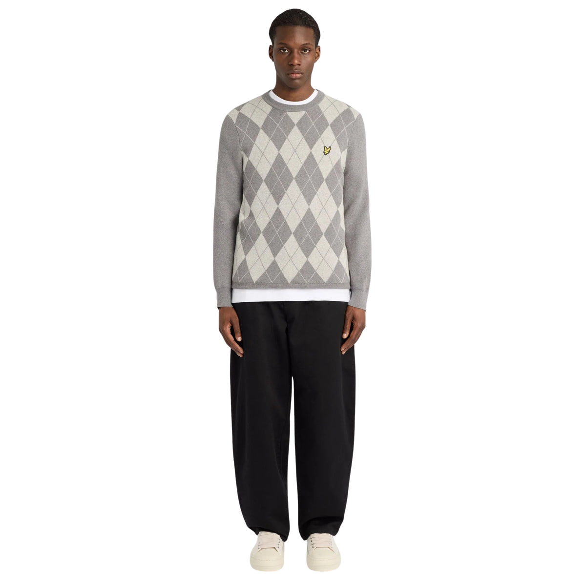 Modelo de corpo inteiro com camisola Lyle & Scott Cotton Merino Argyle cinzenta, calças pretas e sapatilhas cru.