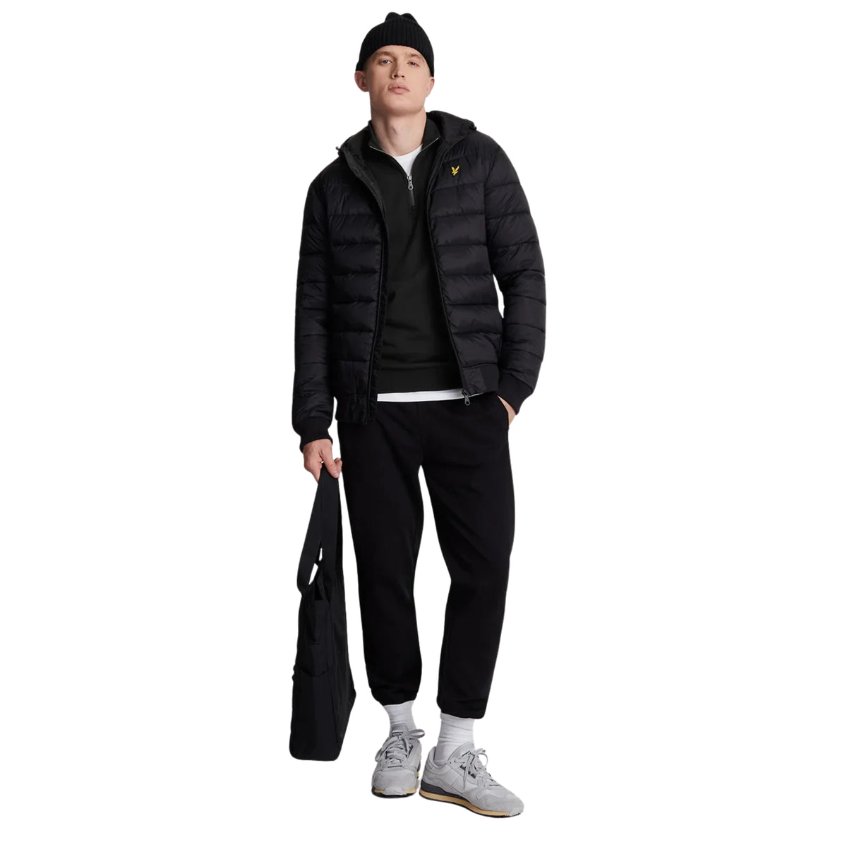 Modelo com sweat Lyle & Scott Loopback Jet Black sob casaco acolchoado aberto, com gorro preto, calças pretas e sapatilhas cinzentas.