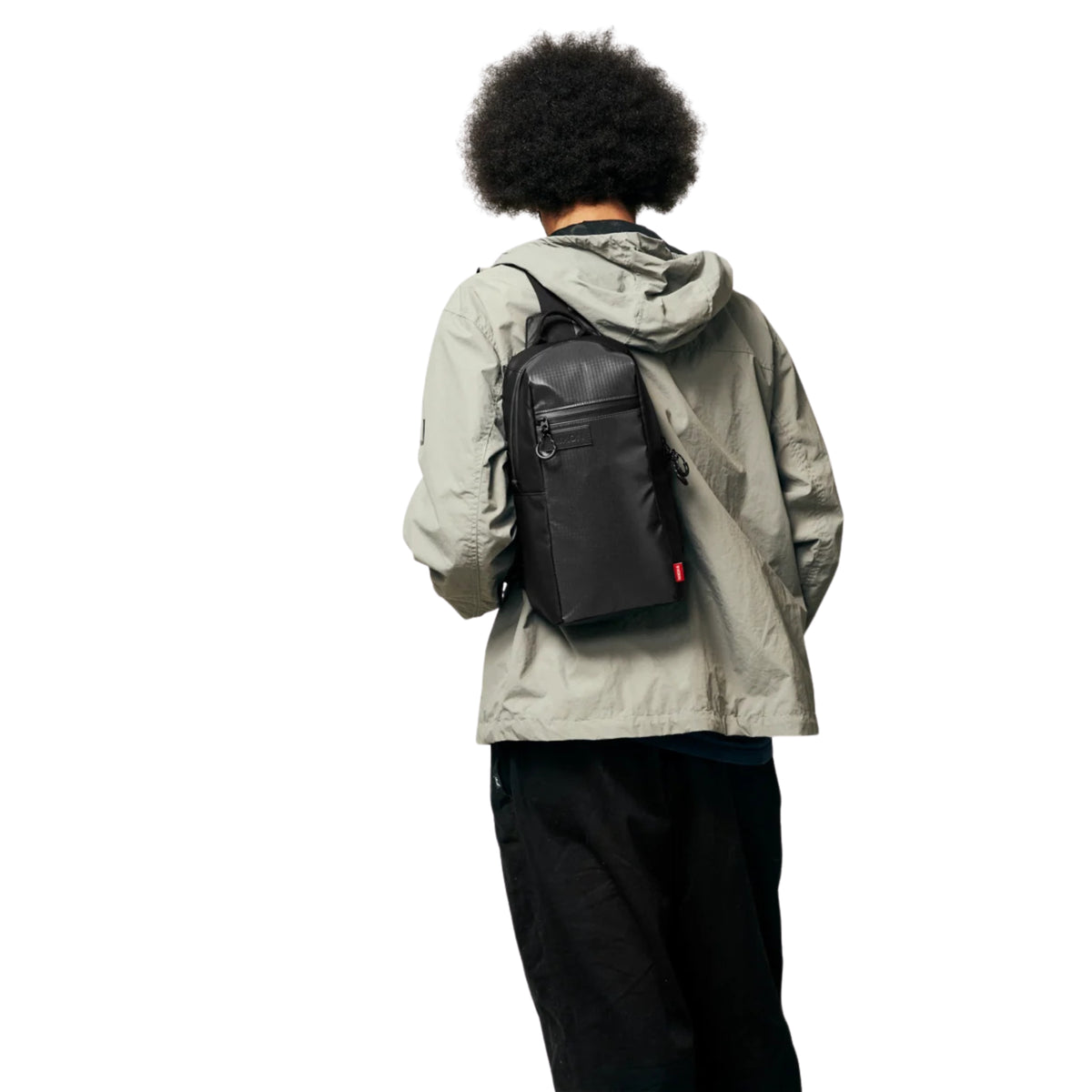 Modelo visto de costas com a bolsa Nixon Syndicate Sling Bag Asphalt, mostrando o formato vertical e o bolso frontal.