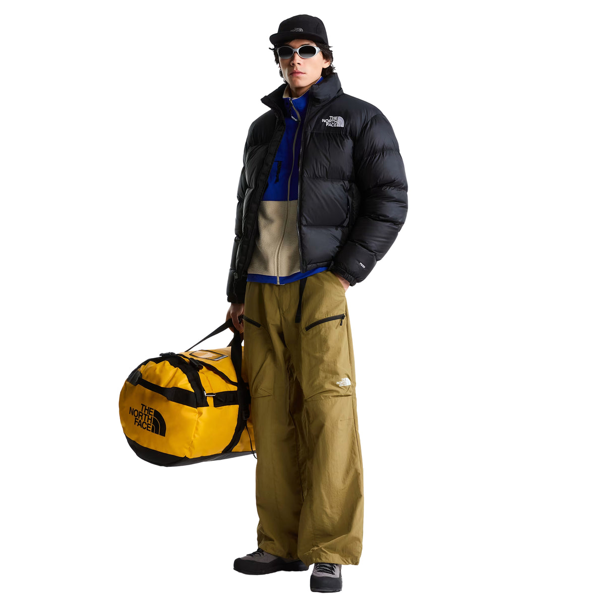 Modelo de corpo inteiro com casaco The North Face 1996 Retro Nuptse preto aberto, calças verde-azeitona e saco de viagem amarelo.
