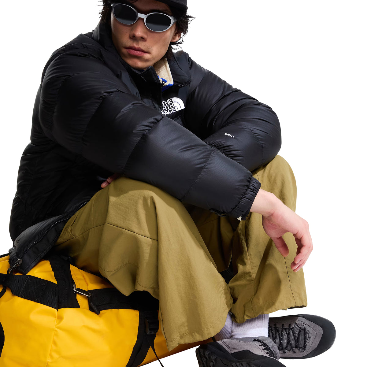 Casaco The North Face 1996 Retro Nuptse preto em modelo sentado sobre um saco amarelo, com destaque para as mangas acolchoadas e o logótipo no peito.