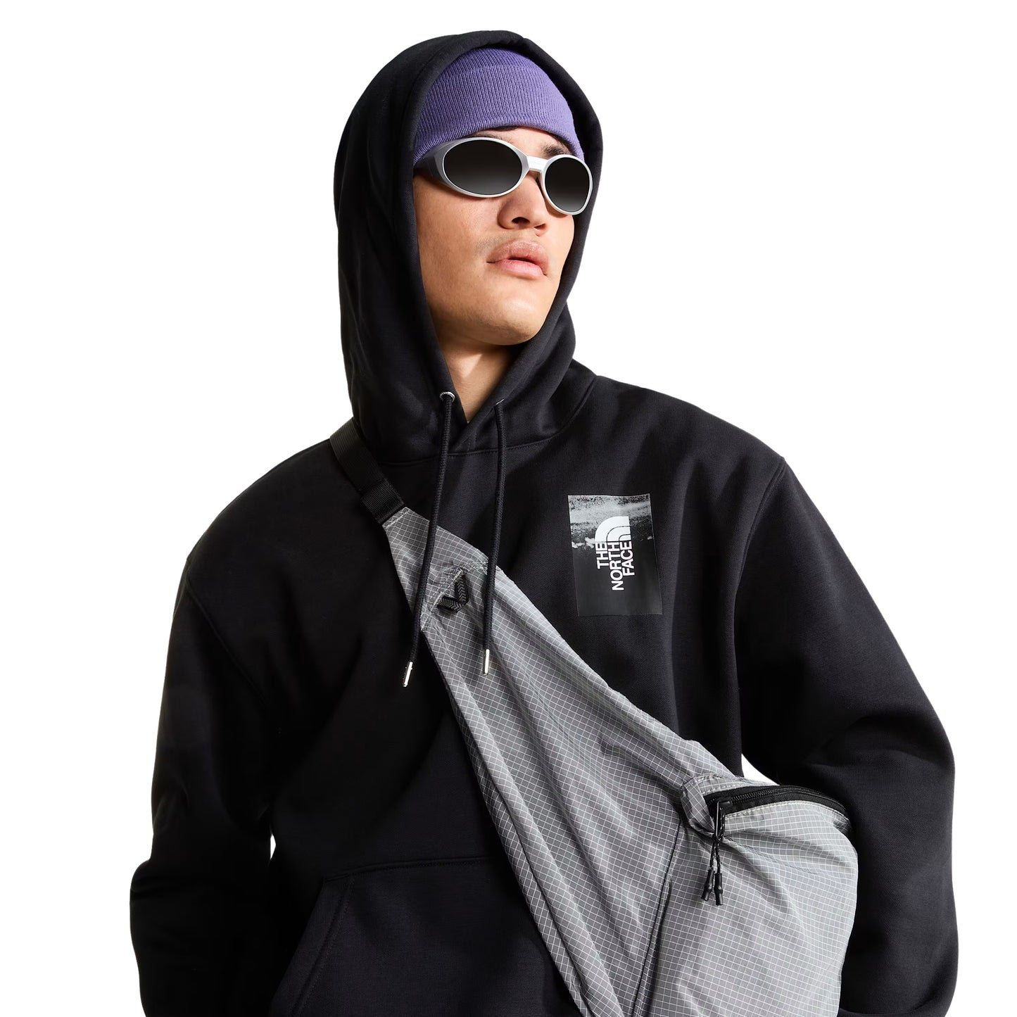 Hoodie The North Face Apex '75 Collage Relaxed em TNF Black — modelo masculino de frente a 3/4, gorro TNF lilás/roxo com headband lilás, óculos de sol ovais prateados, mochila sling TNF cinza em tiracolo, capuz levantado, logo TNF Apex '75 Collage em caixa em serigrafia no peito esquerdo visível, fundo branco.