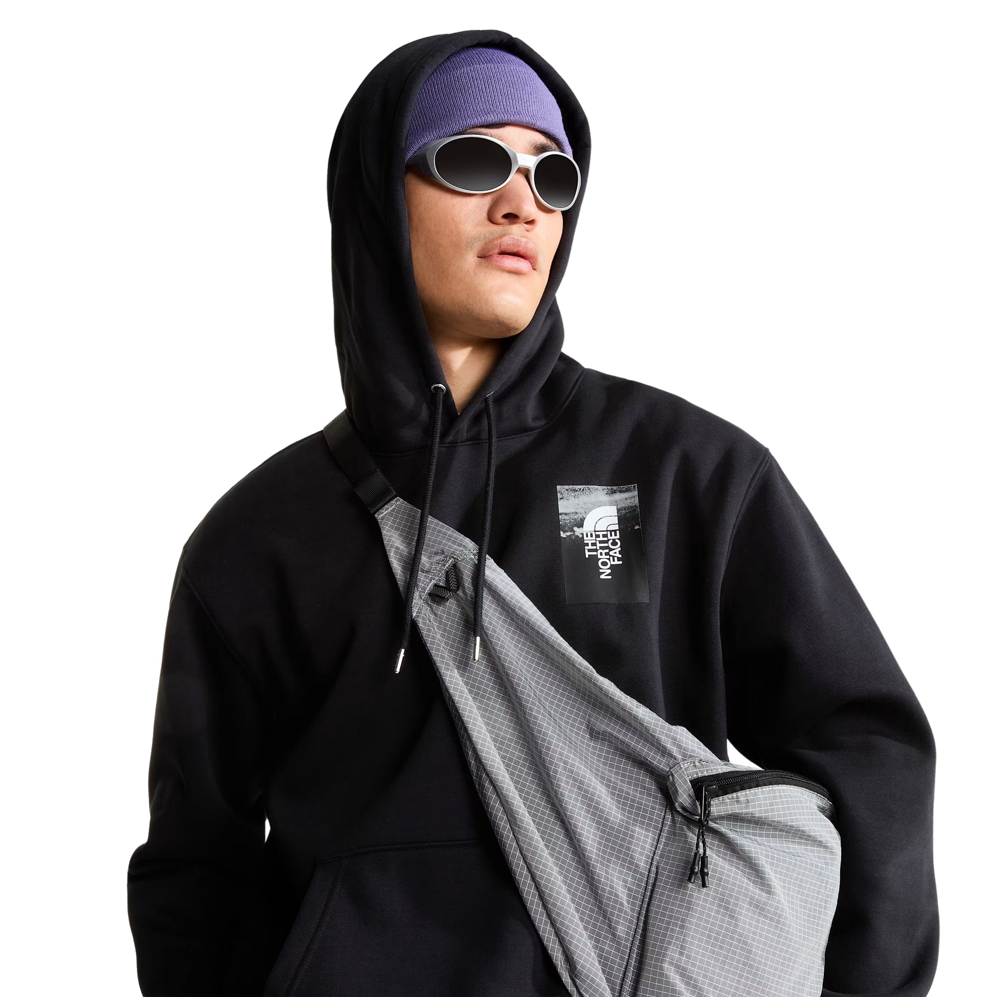 Hoodie The North Face Apex '75 Collage Relaxed em TNF Black — modelo masculino de frente a 3/4, gorro TNF lilás/roxo com headband lilás, óculos de sol ovais prateados, mochila sling TNF cinza em tiracolo, capuz levantado, logo TNF Apex '75 Collage em caixa em serigrafia no peito esquerdo visível, fundo branco.