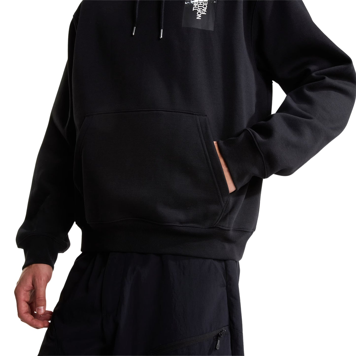Hoodie The North Face Apex '75 Collage Relaxed em TNF Black — detalhe do bolso canguru e bainha canelada de modelo masculino, calças cargo azul-marinho escuro, punho canelado e logo TNF Apex '75 Collage em caixa parcialmente visível no peito, fundo branco.