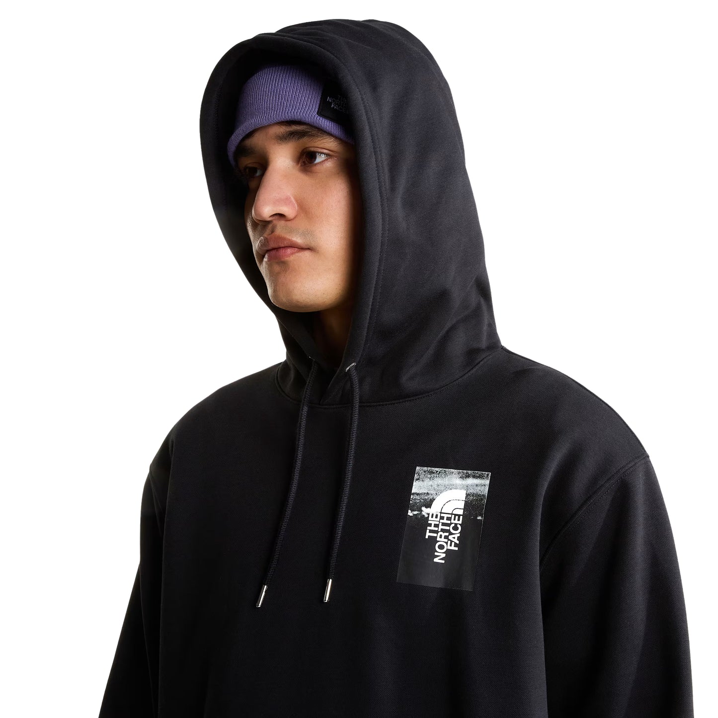 Hoodie The North Face Apex '75 Collage Relaxed em TNF Black — detalhe do peito de modelo masculino com capuz levantado, gorro TNF lilás/roxo, logo TNF Apex '75 Collage em caixa em serigrafia em branco no peito esquerdo e cordão do capuz com ponteiras metálicas visíveis em close-up, fundo branco.