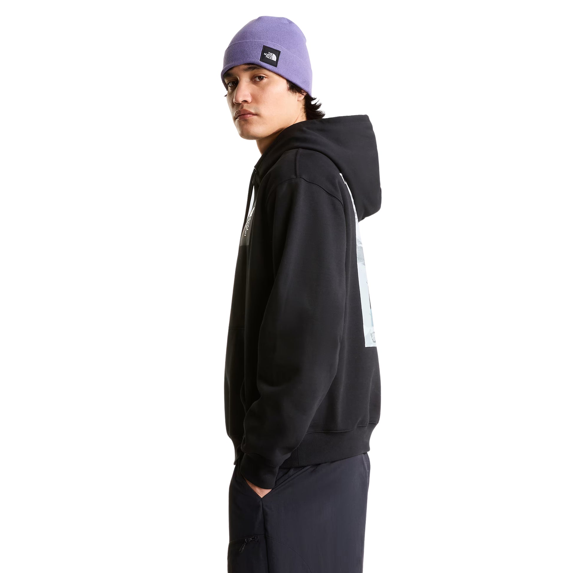 Hoodie The North Face Apex '75 Collage Relaxed em TNF Black — modelo masculino de perfil esquerdo, gorro TNF lilás/roxo com patch TNF quadrado, calças cargo azul-marinho escuro, gráfico collage Apex '75 parcialmente visível nas costas, capuz ajustável visível, fundo branco.