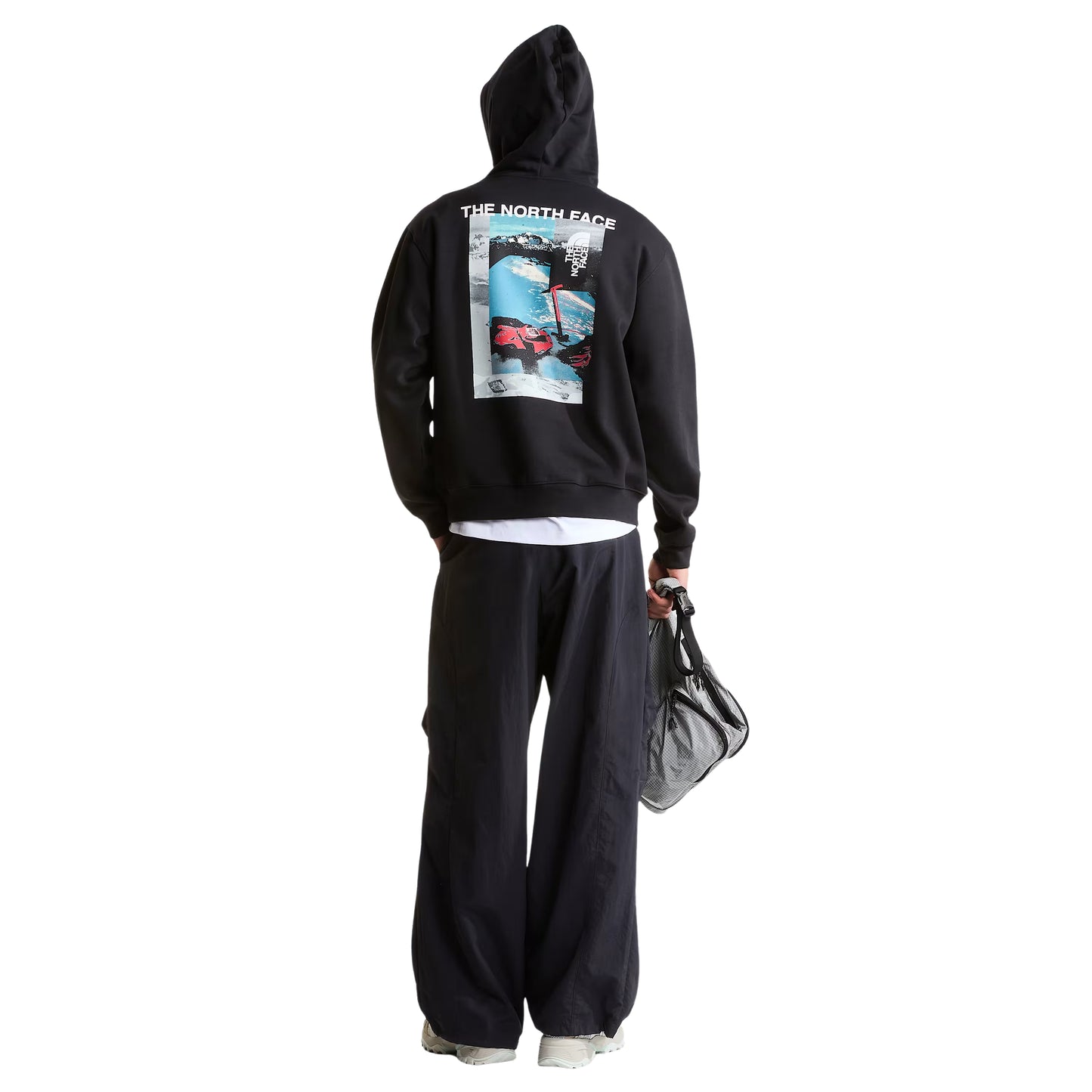 Hoodie The North Face Apex '75 Collage Relaxed em TNF Black — modelo masculino de costas, corpo inteiro, calças largas pretas, sapatilhas brancas, mala de rede preta na mão direita, gráfico collage Apex '75 em caixa com "The North Face" em branco e esquiador em neve azul-glaciar nas costas visíveis, fundo branco.