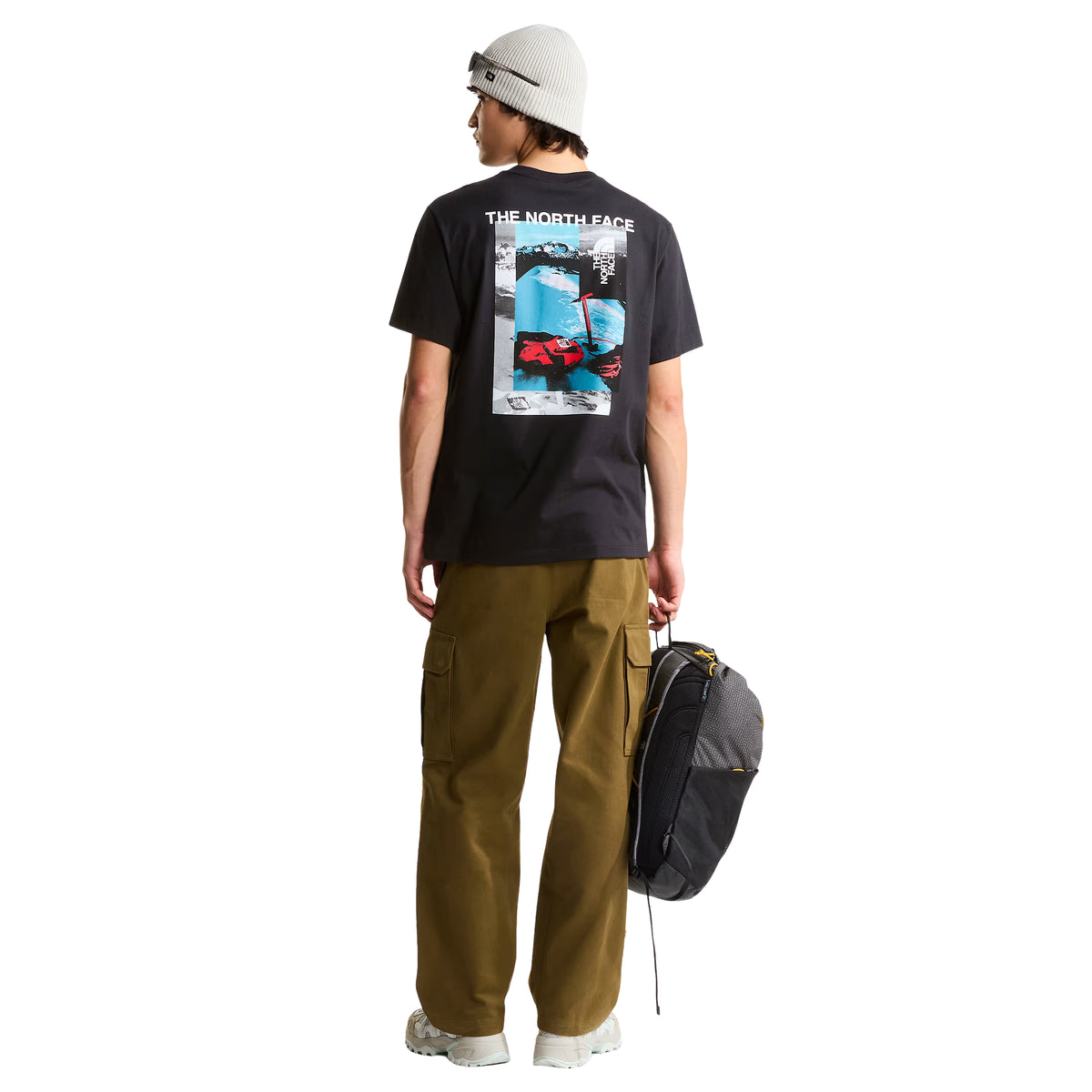 T-shirt de manga curta The North Face Apex '75 Collage em preto — modelo masculino de costas, corpo inteiro, gráfico collage "THE NORTH FACE" em branco, paisagem de montanha nevada em azul/cinzento, luvas e equipamento de ski vermelhos TNF, corte relaxed, calças cargo camel, gorro cinzento, fundo branco.