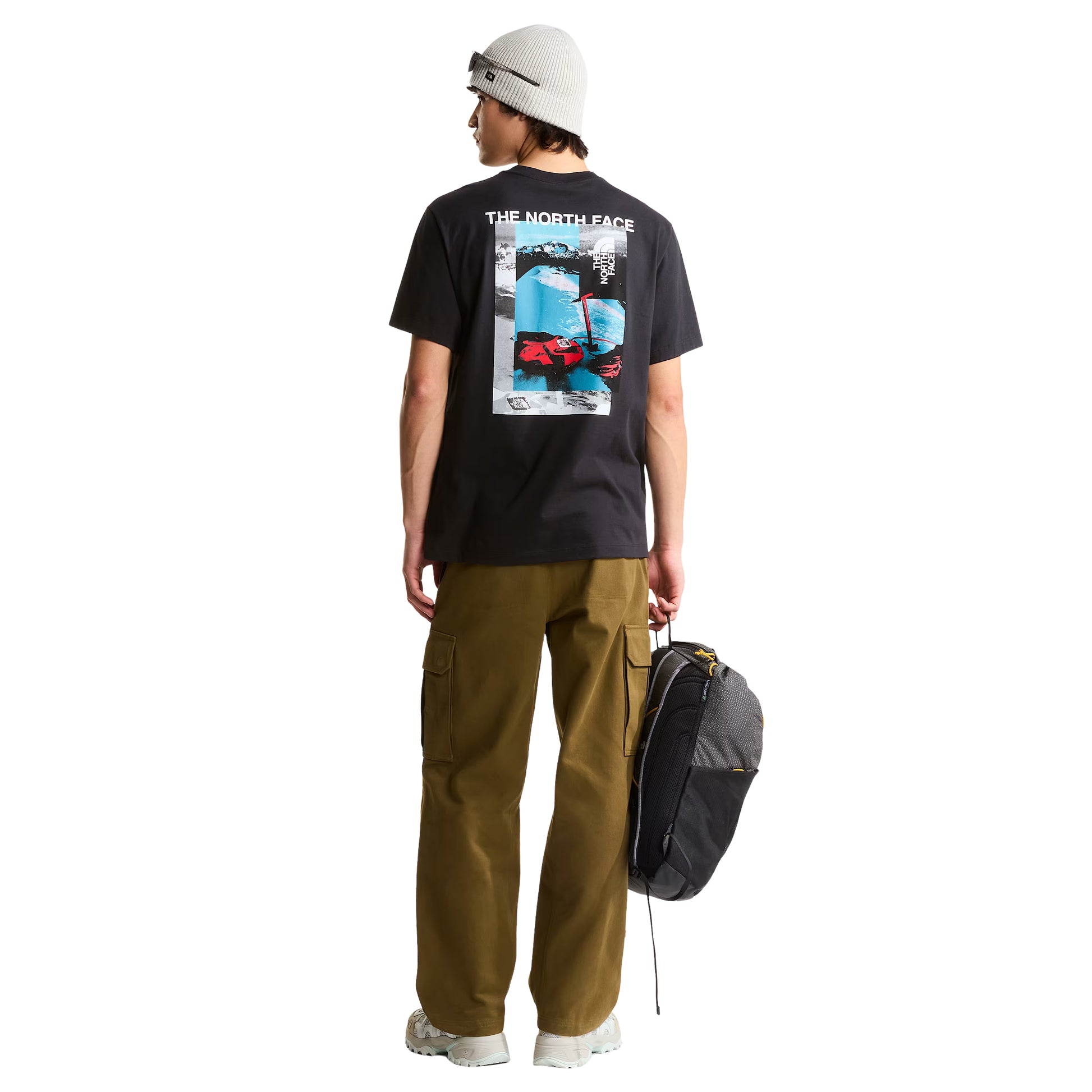 T-shirt de manga curta The North Face Apex '75 Collage em preto — modelo masculino de costas, corpo inteiro, gráfico collage "THE NORTH FACE" em branco, paisagem de montanha nevada em azul/cinzento, luvas e equipamento de ski vermelhos TNF, corte relaxed, calças cargo camel, gorro cinzento, fundo branco.