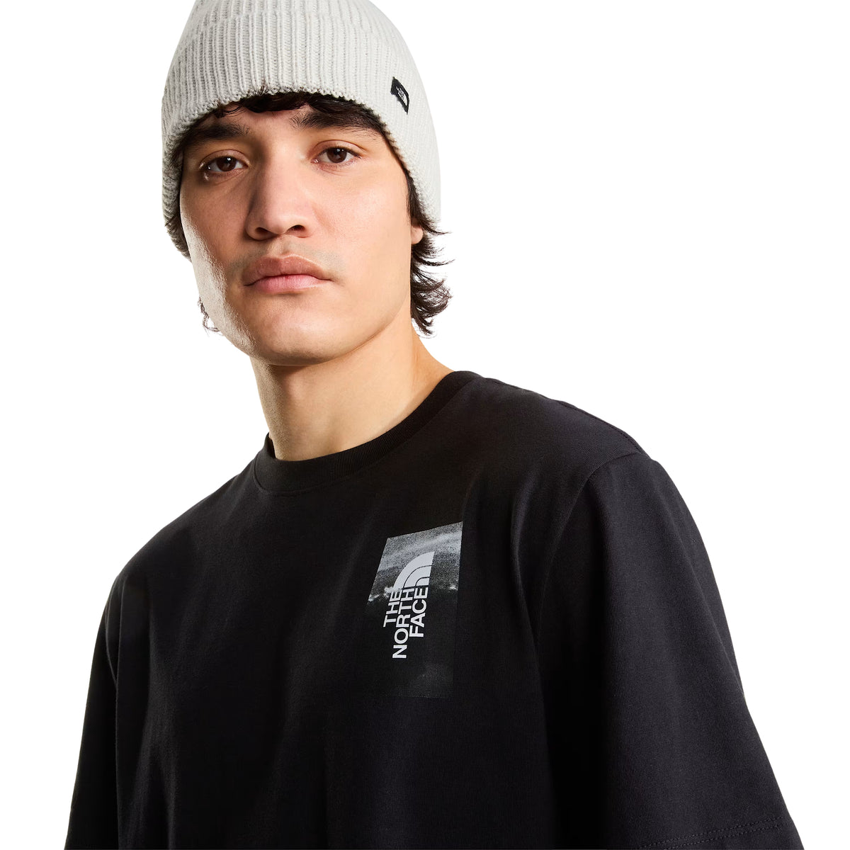 T-shirt de manga curta The North Face Apex '75 Collage em preto — modelo masculino de frente, meio corpo, pormenor do peito esquerdo com logótipo The North Face em retângulo fotográfico sobre fundo preto, gola redonda canelada, gorro cinzento, fundo branco.