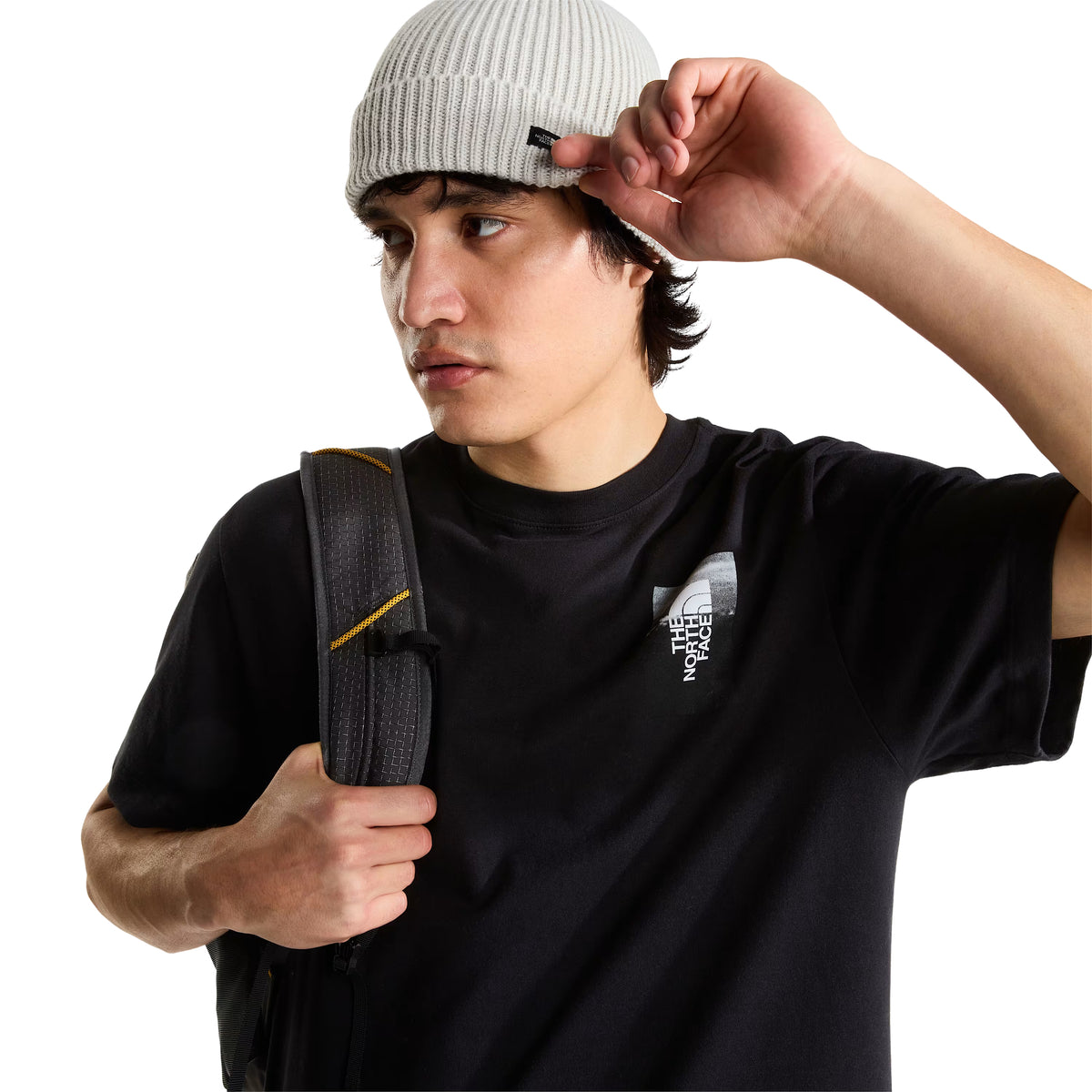 T-shirt de manga curta The North Face Apex '75 Collage em preto — modelo masculino de frente, meio corpo, logótipo The North Face em retângulo fotográfico cinzento/branco no peito esquerdo, gola redonda canelada, corte relaxed, mochila cinzenta escura com alça amarela a tiracolo, gorro cinzento claro The North Face, fundo branco.