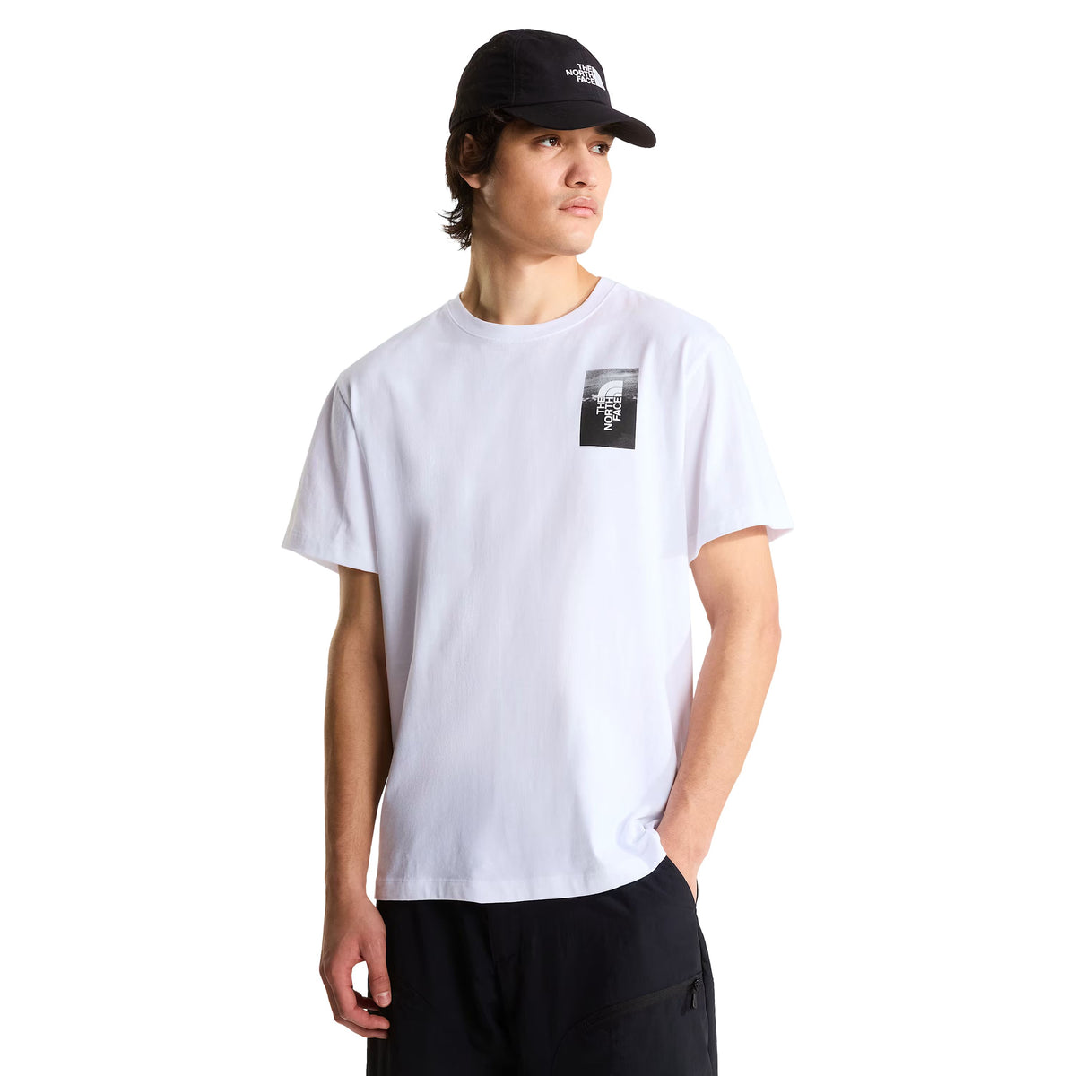 T-shirt de manga curta The North Face Apex '75 Collage em branco — modelo masculino de frente, meio corpo, t-shirt branca com logótipo The North Face em retângulo preto e branco no peito esquerdo, gola redonda canelada, corte relaxed, calças pretas largas, boné preto The North Face, fundo branco