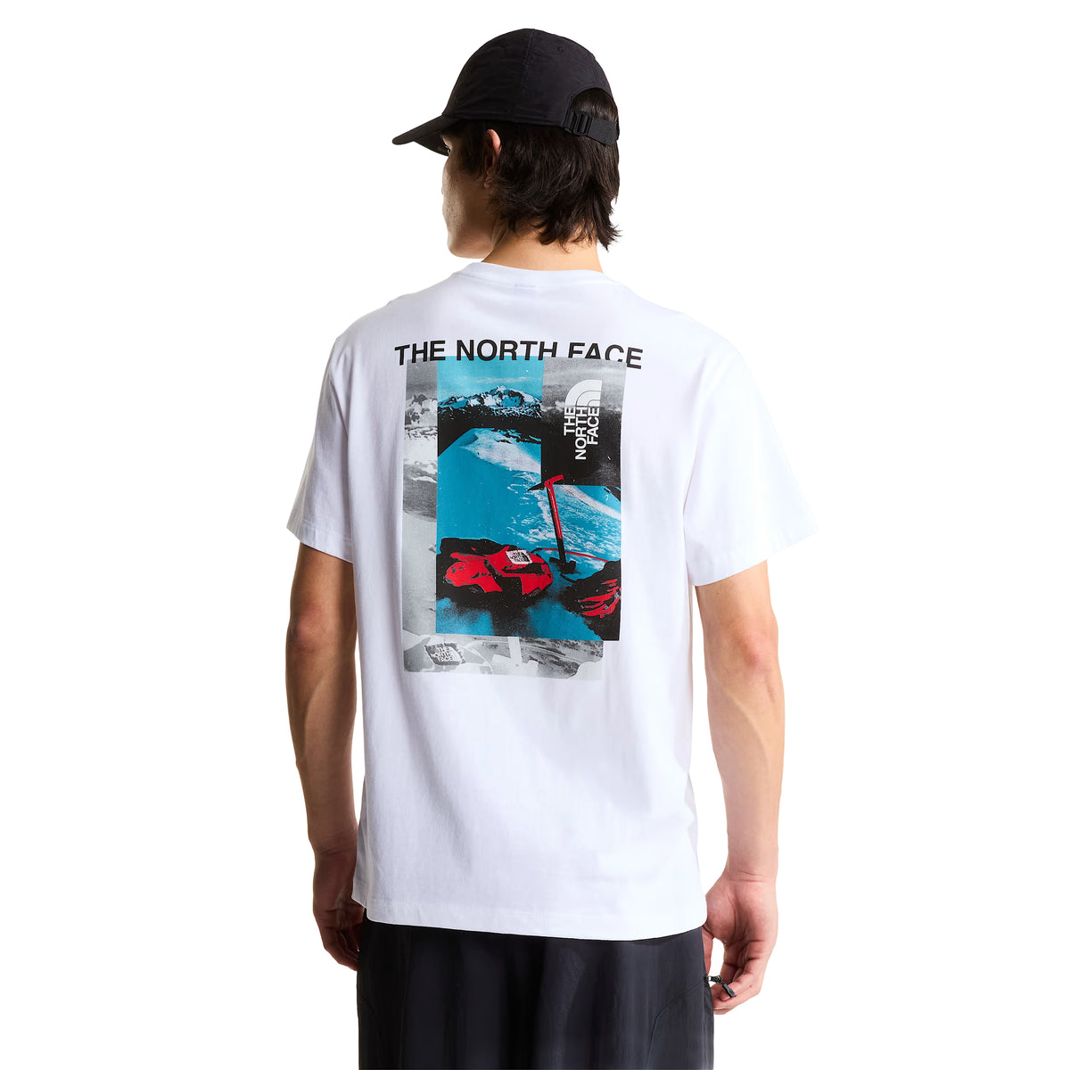 T-shirt de manga curta The North Face Apex '75 Collage em branco — modelo masculino de costas, corpo inteiro, gráfico collage com "THE NORTH FACE" em preto, paisagem de montanha nevada em azul/cinzento, luvas e equipamento de ski vermelho TNF, corte relaxed, boné preto, calças pretas, fundo branco.