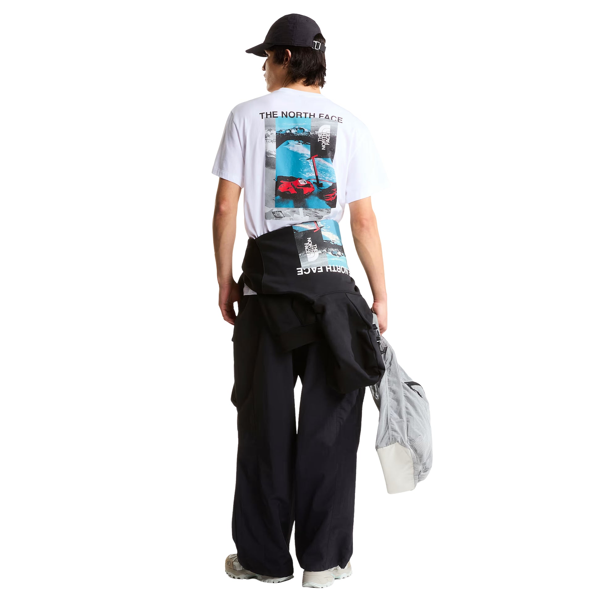 T-shirt de manga curta The North Face Apex '75 Collage em branco — modelo masculino de costas, corpo inteiro, gráfico collage Apex '75 com "THE NORTH FACE" em preto, paisagem nevada em azul/cinzento e luvas de ski vermelhas TNF, calças cargo pretas largas com casaco amarrado à cintura, boné preto, sapatilhas bege, fundo branco.
