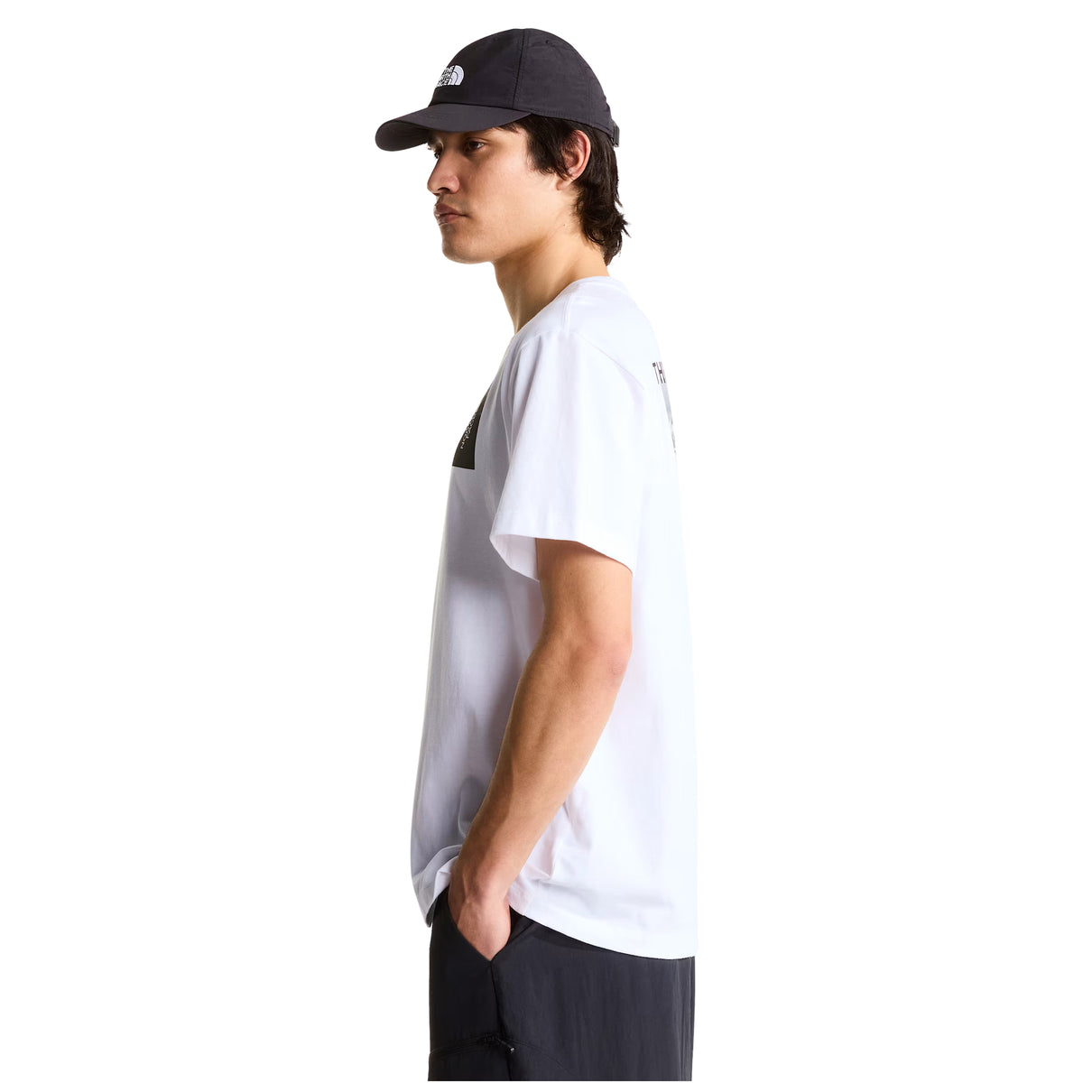 T-shirt de manga curta The North Face Apex '75 Collage em branco — modelo masculino de perfil direito, meio corpo, t-shirt branca com logótipo The North Face em retângulo preto e branco visível no peito, gola redonda canelada, corte relaxed, calças pretas, boné preto The North Face, fundo branco.