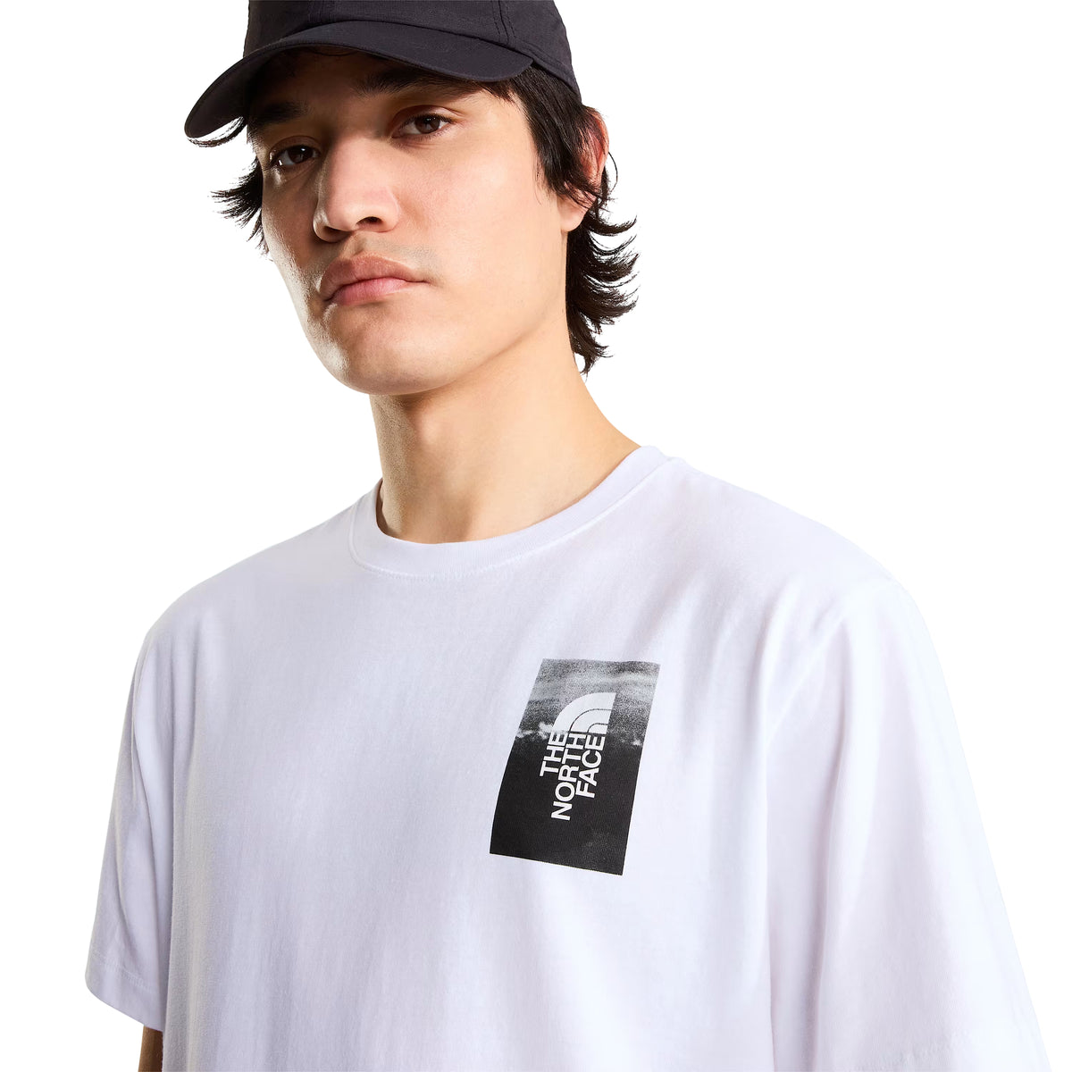 T-shirt de manga curta The North Face Apex '75 Collage em branco — modelo masculino de frente, pormenor do peito esquerdo, logótipo The North Face em retângulo preto e branco com montanha nevada no topo, gola redonda canelada, boné preto, fundo branco.