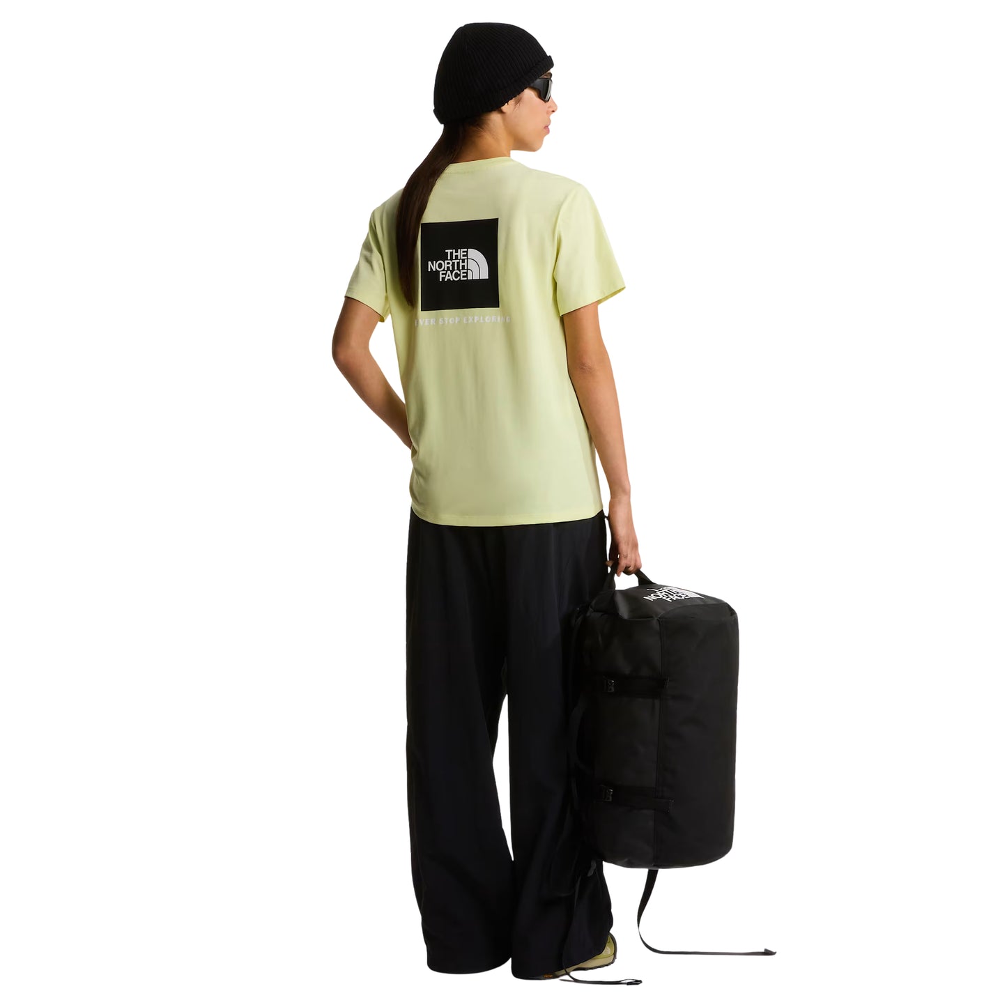 Saco de viagem The North Face Base Camp Duffel XS em TNF Black — modelo feminino de costas, t-shirt amarelo lima com logo TNF em caixa preta, calças largas pretas, gorro preto e óculos de sol escuros, saco transportado pela alça de mão com logo TNF em branco visível de lado, fundo branco.
