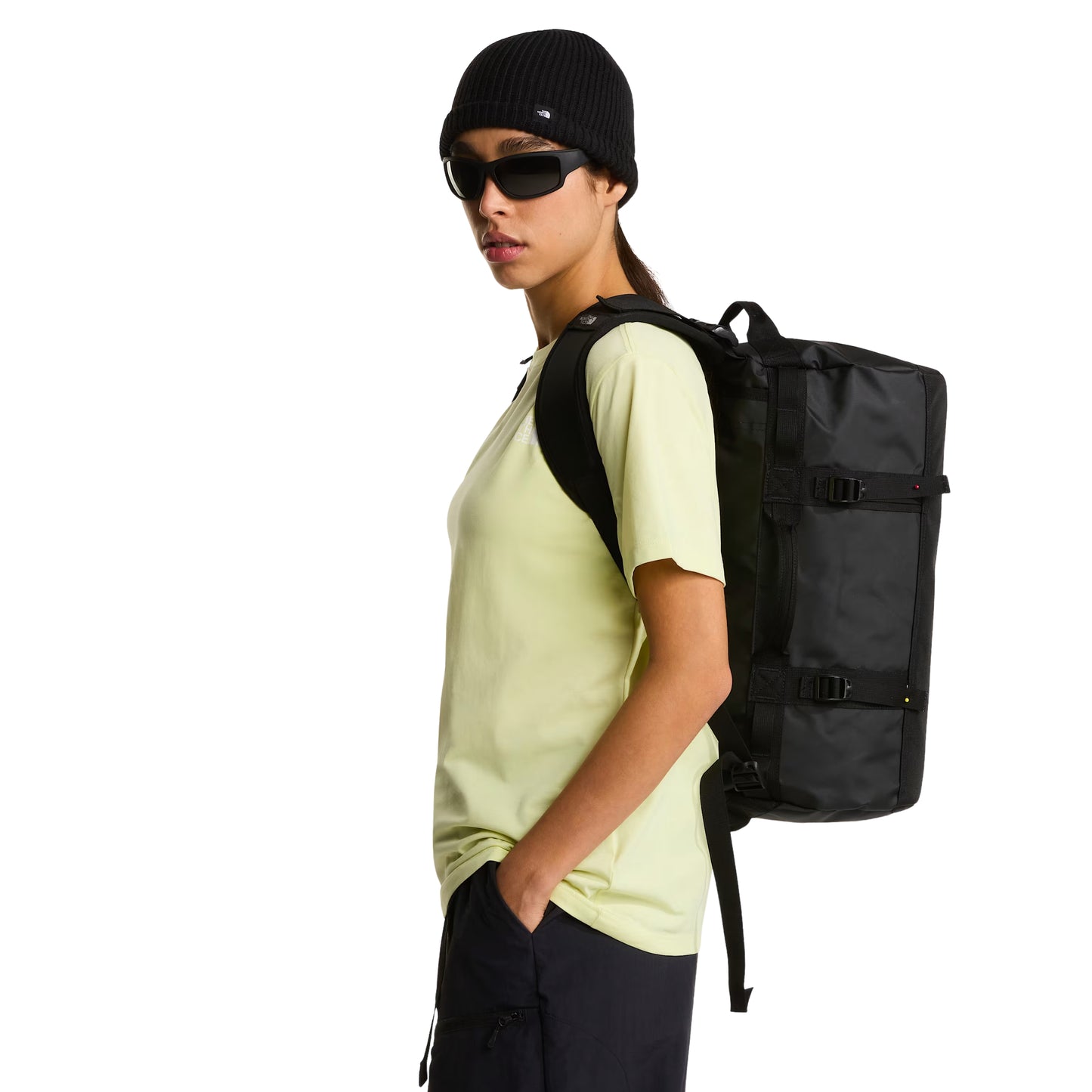 Saco de viagem The North Face Base Camp Duffel XS em TNF Black — modelo feminino de perfil, t-shirt amarelo lima, gorro TNF preto e óculos de sol escuros, saco transportado às costas com alças de mochila pretas ajustadas, fundo branco.