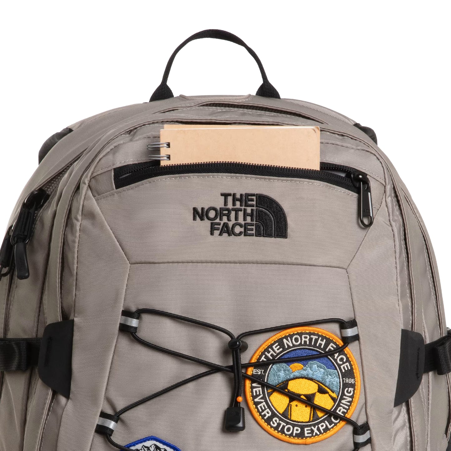 Mochila The North Face Borealis Classic Patch em Stone Slab / TNF Black — detalhe do topo com bolso zip aberto e caderno bege, logo The North Face bordado em preto e patch circular "Never Stop Exploring" amarelo/laranja visíveis.