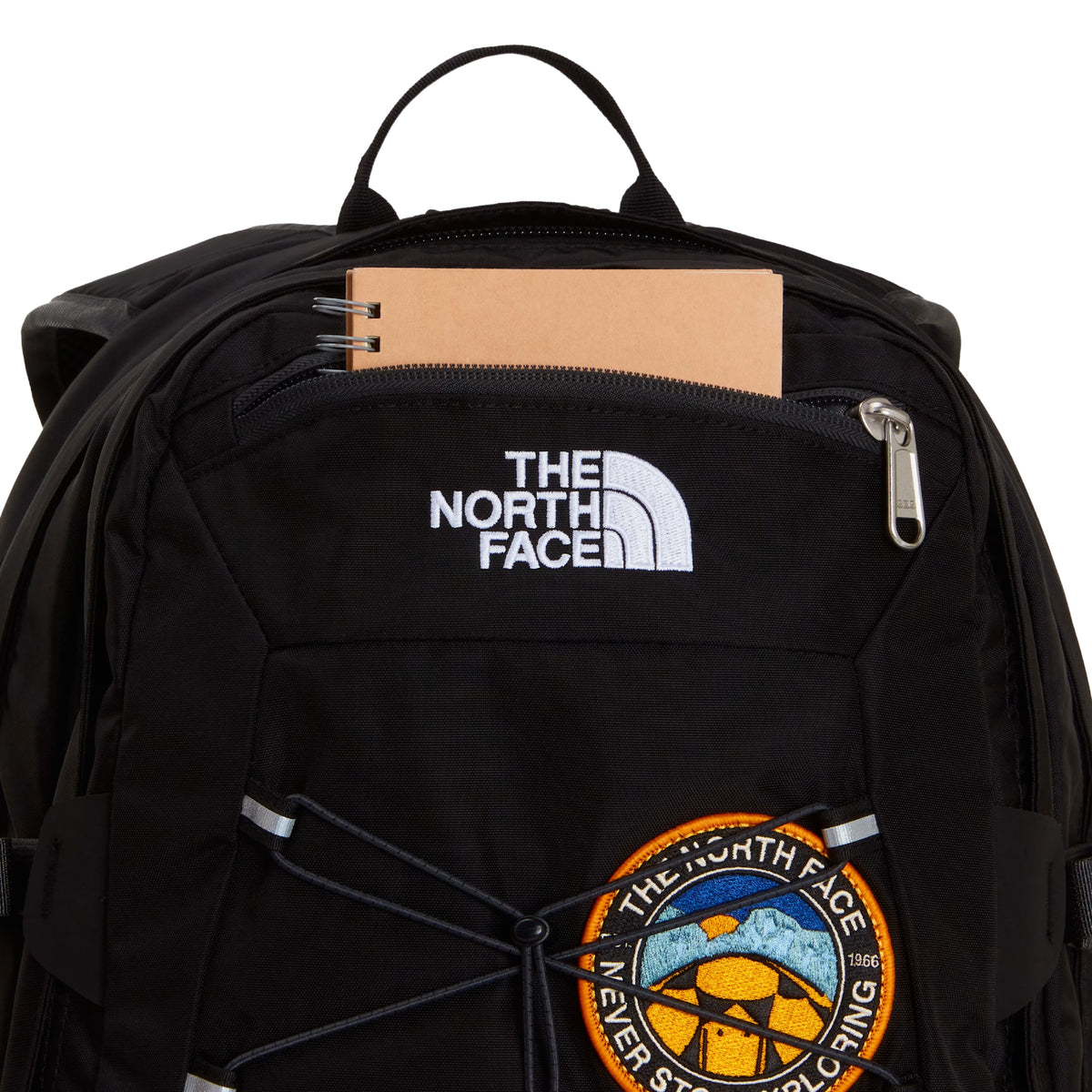 Mochila The North Face Borealis Classic Patch em TNF Black — detalhe do topo com bolso zip aberto e caderno bege, logo The North Face bordado em branco e patch circular "Never Stop Exploring" laranja/amarelo visíveis.